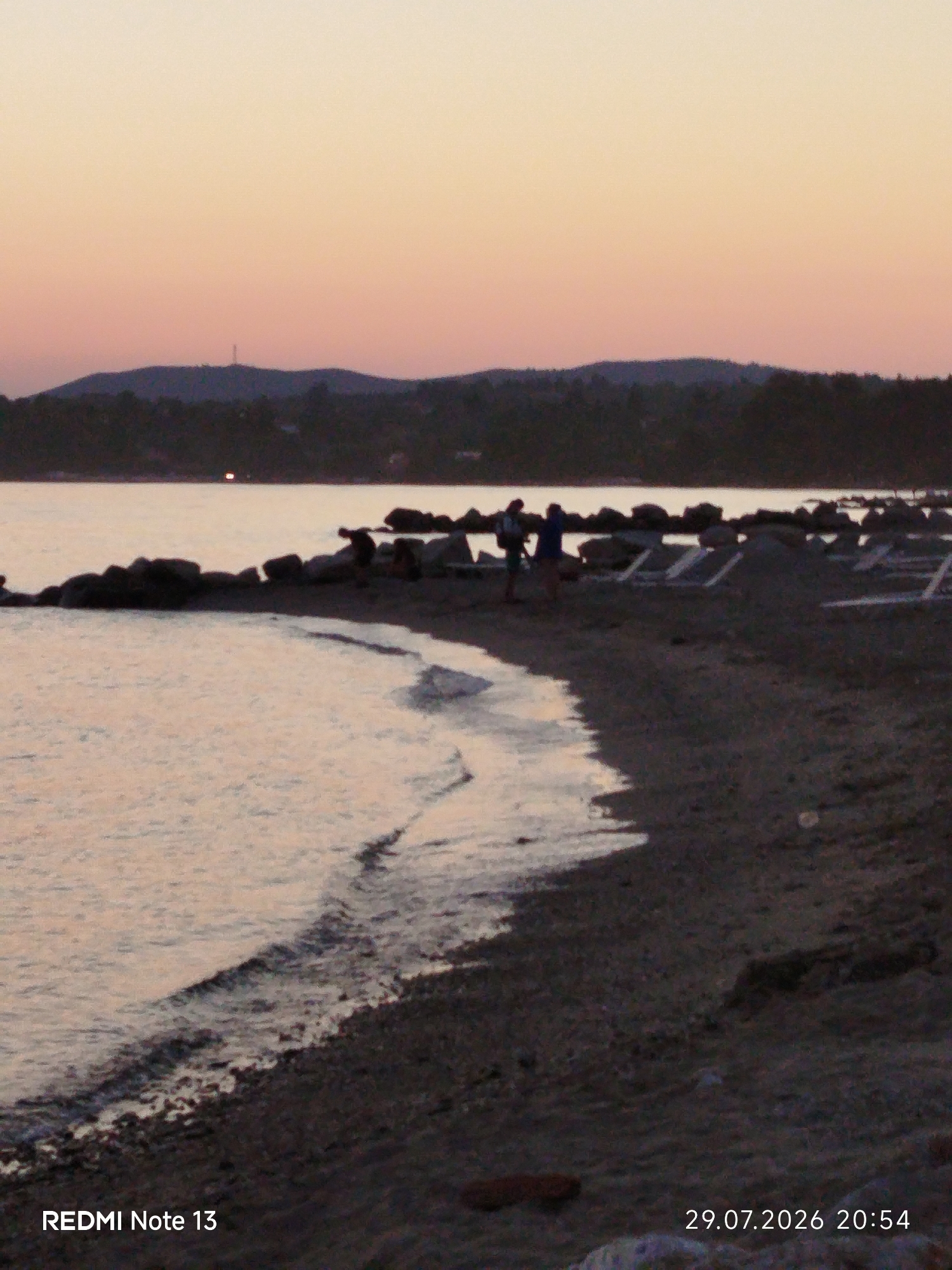The image depicts a serene coastal scene at dusk in the Sithonia Municipal Unit, Greece. A gentle wave laps the pebbly shore, with a line of dark rocks separating the water from the beach. Several beach chairs are arranged on the sand, some appearing to be stacked. In the background, a dark, tree-covered hill is silhouetted against a soft, peach-colored sky. A distant transmission tower is visible on the hilltop. Two figures are standing on the beach, their forms indistinct in the low light. The bottom left of the image displays "REDMI Note 13" and the bottom right shows "29.07.2026 20:54".