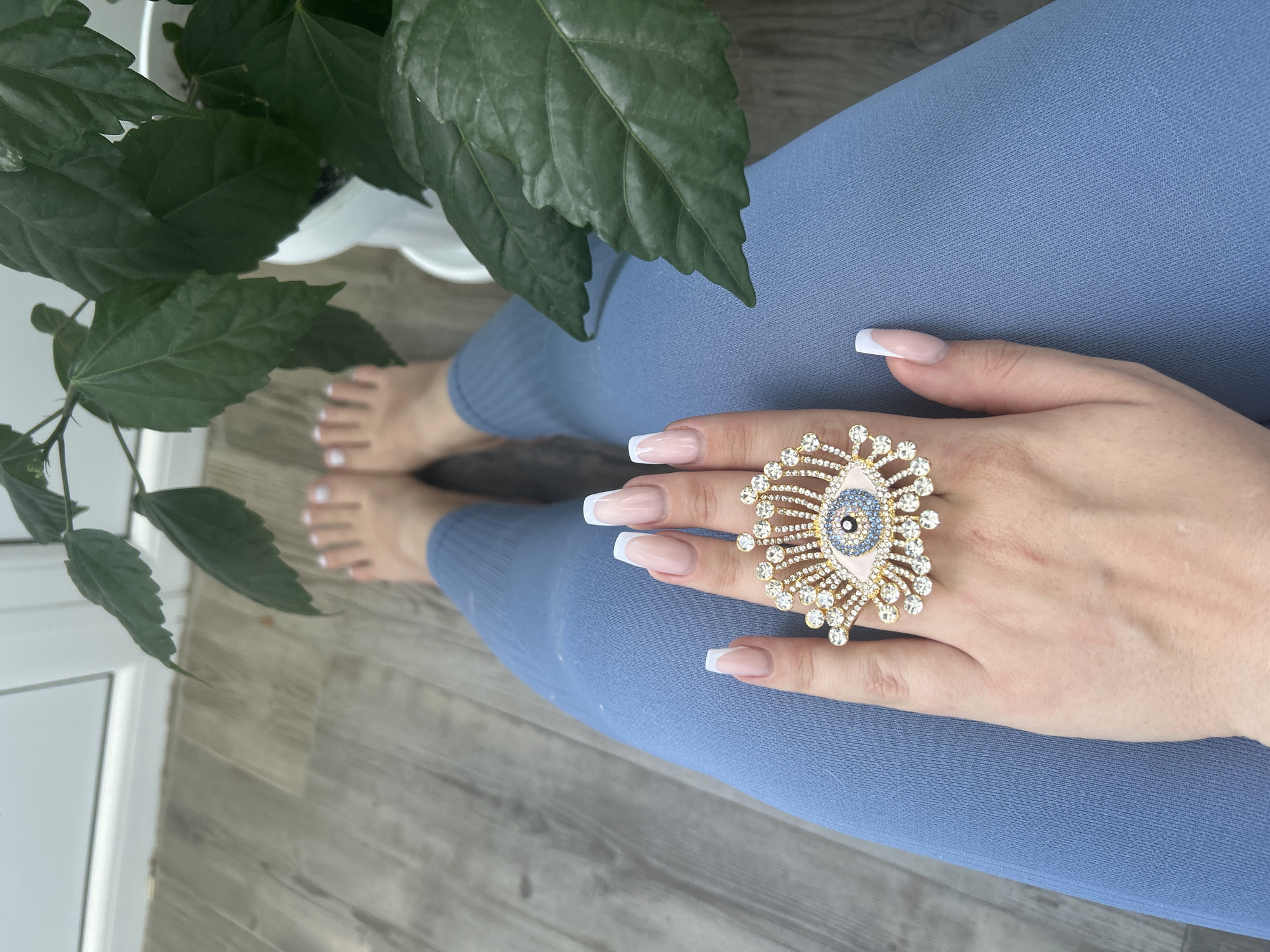 An overhead, medium shot captures a person's legs and hands, adorned with a large, bejeweled evil eye ring, set against a neutral, wooden floor. The person is wearing blue leggings, and their feet, with white-painted toenails, are visible at the top of the frame. A large potted plant with dark green leaves is situated to the right, partially obscuring the background. The scene appears to be indoors, with a wall and window visible in the upper portion of the image. The image does not provide any information about the city of Odesa, Ukraine.