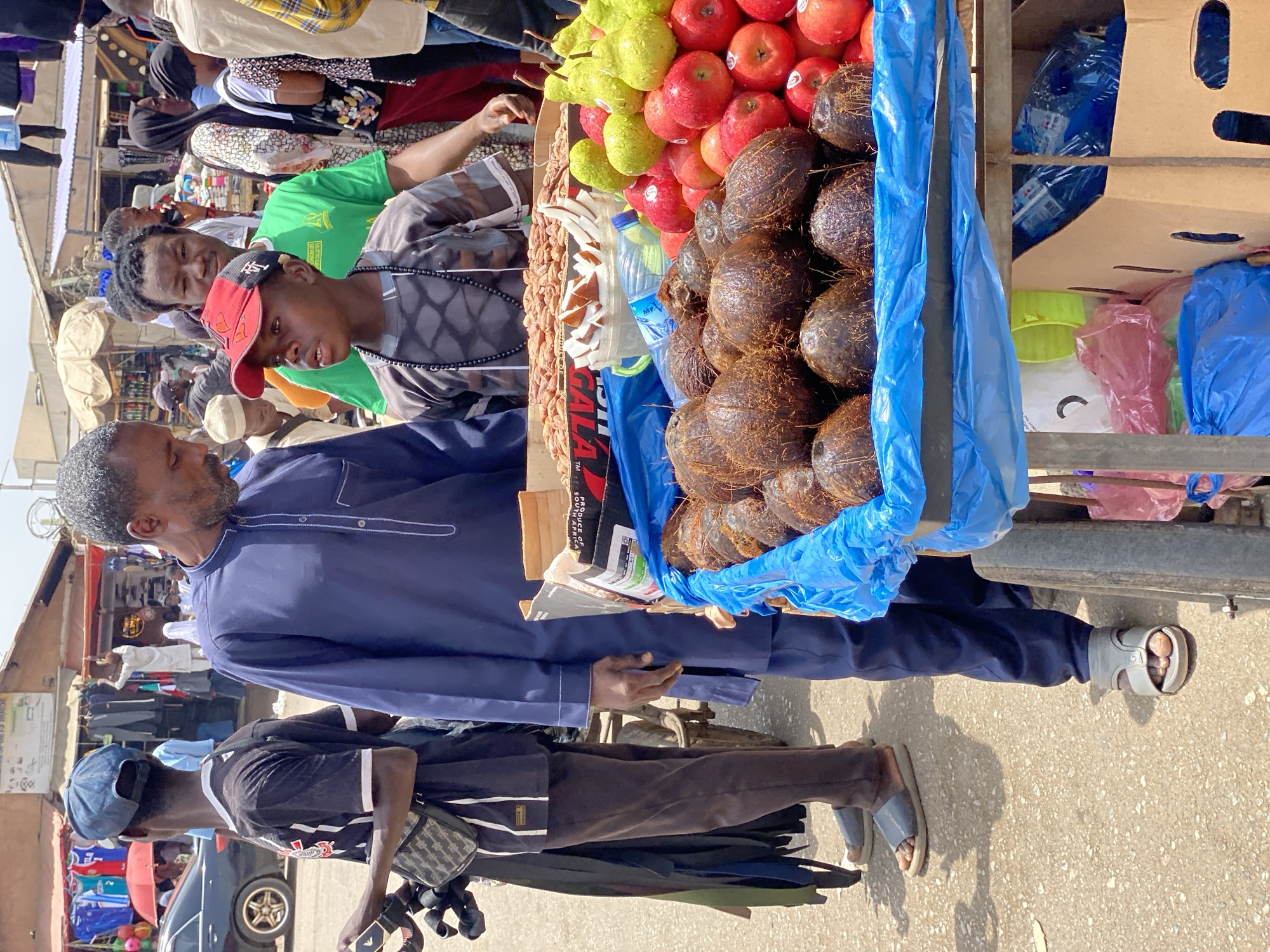 In Maiburiji, Nigeria, a street scene unfolds. A man in a blue shirt stands beside a cart filled with goods. On the cart are red apples, green pears, and a pile of brown coconuts, all contained within a blue plastic liner. A water bottle is also visible. Several other people stand near the cart. One is wearing a green shirt and another, a red baseball cap. Buildings are visible in the background and there's a car. The ground is light-colored and appears to be a street or paved area.