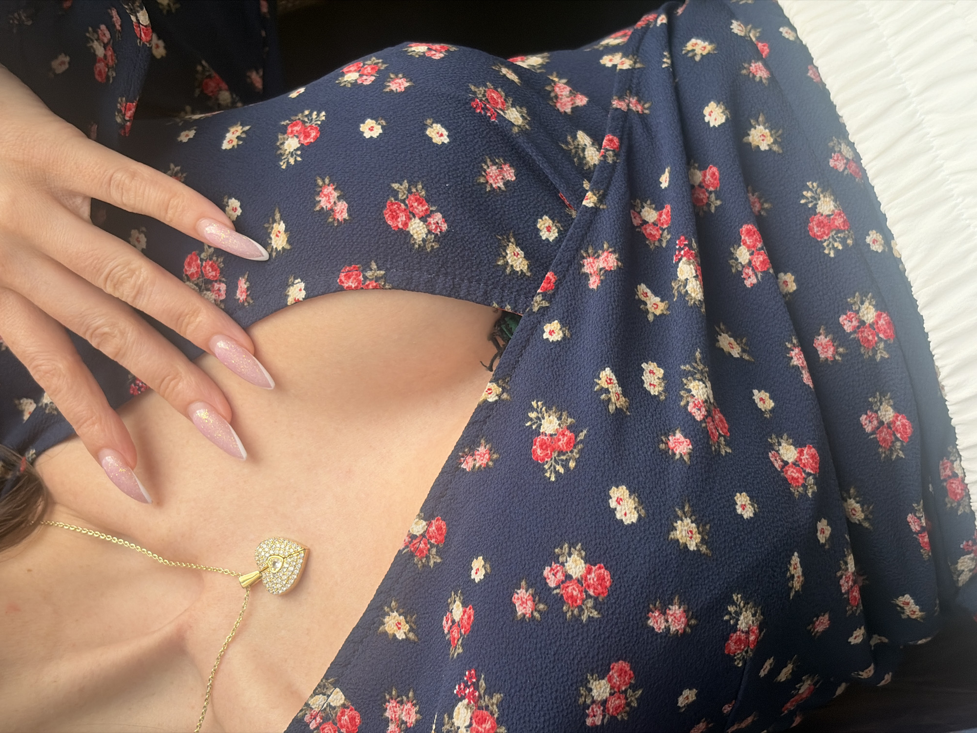 The image captures a close-up, high-angle view of a person, likely female, from the chest upwards, reclining or lying down. The subject is wearing a dark navy blue dress or top patterned with small floral motifs in shades of red, pink, and off-white, on a textured fabric.
A manicured hand with long, almond-shaped fingernails rests gently on the chest area. The nails are styled with a glittery light pink base and white tips, resembling a French manicure. A delicate gold chain necklace is visible, featuring a prominent heart-shaped pendant embellished with numerous small, sparkling clear stones and a larger central clear stone set within a smaller heart outline.
A small portion of what appears to be a dark tattoo is subtly visible in the cleavage area, partly obscured by the dress. To the right of the frame, a white, ruffled or textured fabric, possibly a bedsheet or cushion, is present. The lighting is soft, highlighting the intricate details of the nails, jewelry, and dress pattern. The overall scene focuses on personal style and accessories in a relaxed, intimate setting. No specific location cues related to Saint Petersburg, Russia, are visible within the image.