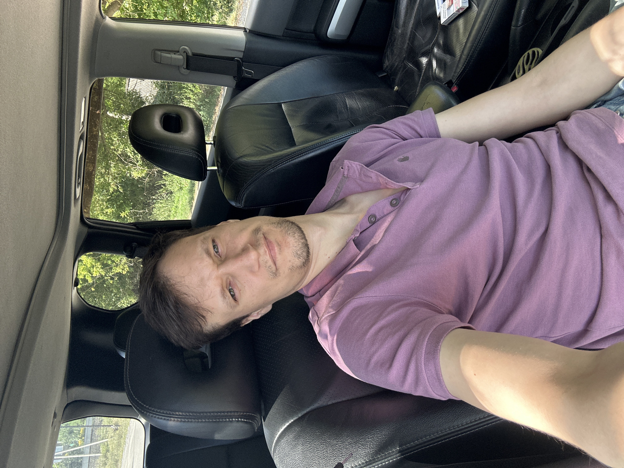 A man with short brown hair and a light beard is sitting in the driver's seat of a car. He is wearing a pink polo shirt and looking at the camera with a slight smile. The car's interior is visible, with black leather seats and a sunroof. Outside the car, trees and a fence can be seen, suggesting a rural or suburban setting, possibly on the outskirts of Makiv, Ukraine. The lighting indicates it is daytime, with sunlight streaming into the car.