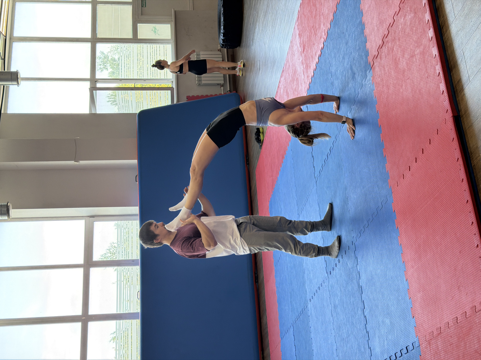 In a gymnastics hall, possibly located in Chaikovsky, Russia, a man in a maroon and white t-shirt and grey sweatpants assists a woman performing a gymnastic bridge pose. The woman, wearing a grey sports bra and black shorts, balances on her hands while her legs are arched backward, her feet being held by the man. Another woman, dressed in a black sports bra and shorts, stands in the background near a large window. The floor is covered with blue and red interlocking mats. A large blue mat is visible in the background, along with a large black tire and gym equipment. The environment suggests a dedicated space for athletic training.