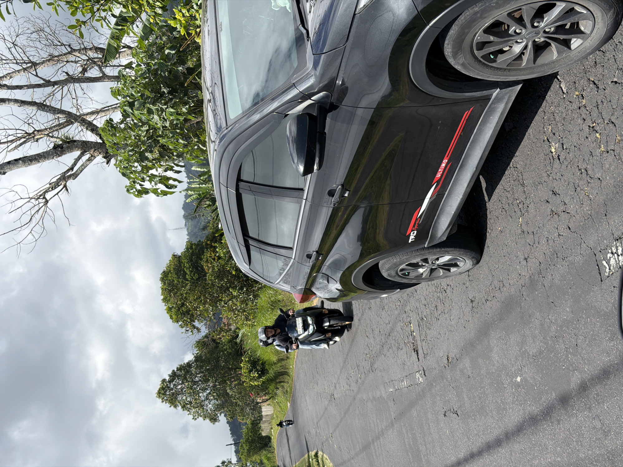 In Kintamani, Indonesia, a black SUV is parked on an asphalt road, its front passenger side visible. The SUV has a sticker with red and white racing stripes on its lower body, reading "TRD Sportivo ULTIMO." The road is cracked and uneven, with shadows cast from overhead trees. Two individuals on a motorcycle, wearing helmets, ride away from the viewer on the same road, further down. A second, smaller motorcycle is visible in the distance. The environment is lush with green trees and vegetation, under a cloudy sky with patches of blue. The terrain appears hilly, with a distant mountain visible in the background.
