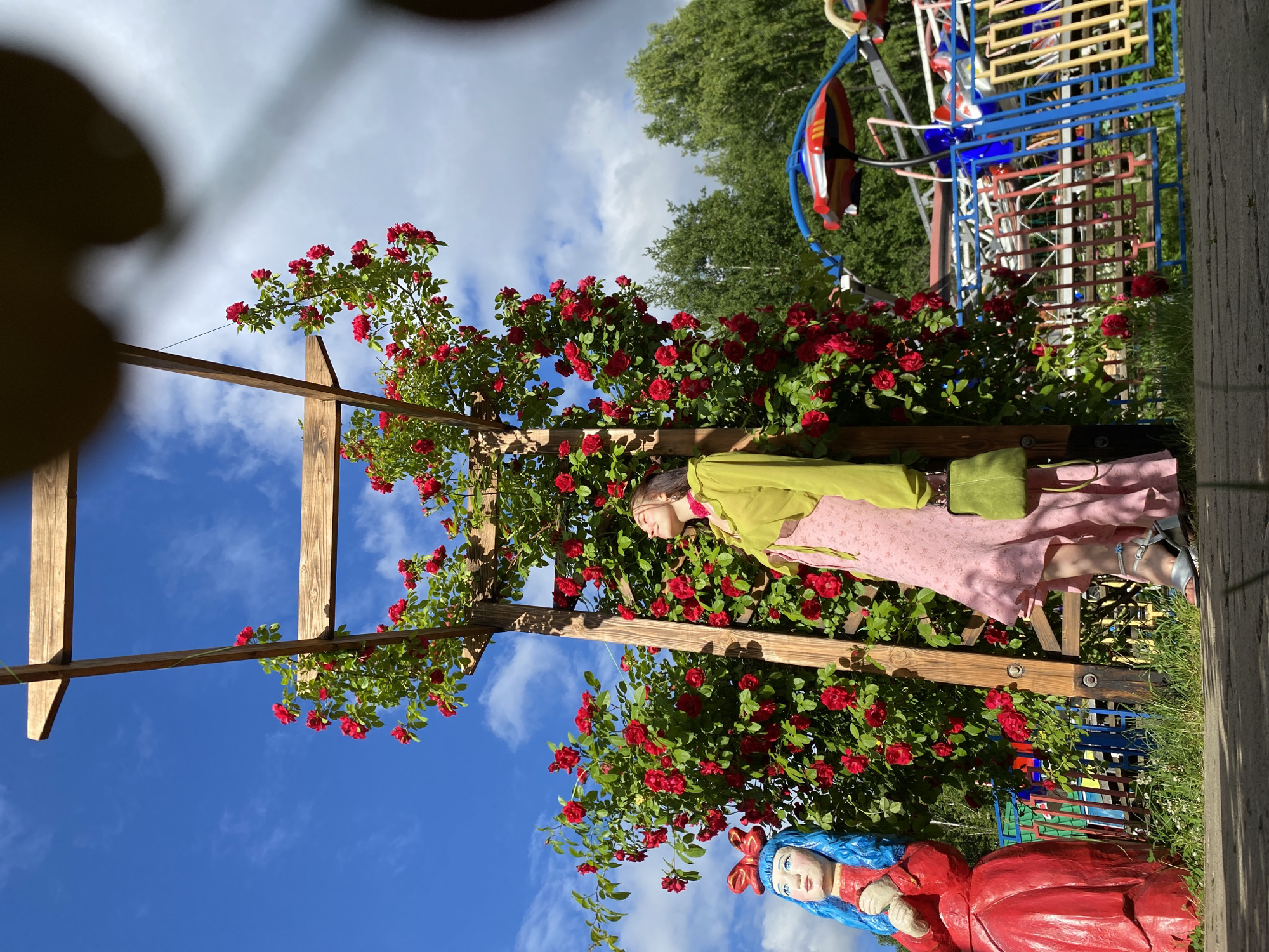 A vertical shot depicts a garden scene with a person and decorative figures among blooming red roses. The person, facing away from the camera and slightly to the right, is wearing a light green jacket over a pink dress and silver sandals. They are leaning against a wooden pergola structure adorned with climbing roses. To the lower right, a decorative figure with bright blue hair and a red dress is visible, with a red bow on its head. The garden is lush with green foliage and vibrant red roses. In the background, a colorful abstract sculpture or playground structure can be seen, along with mature green trees under a bright blue sky with scattered white clouds. The overall impression is a sunny day, likely late morning or afternoon, in a well-maintained outdoor garden space. The scene has a whimsical and serene feel. No visible text is present in the image. The location is Chaikovsky, Russia.