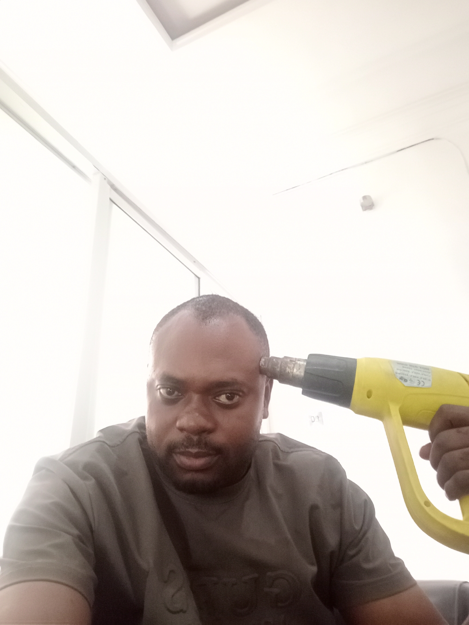 An adult male with dark skin and a serious or neutral expression is depicted in a close-up indoor shot. He has short, dark hair with a receding hairline and is wearing a dark, possibly grey, short-sleeved t-shirt. The man is holding a yellow and black heat gun to his right temple with his right hand, mimicking the gesture of holding a firearm to his head.
The background is uniformly bright white, suggesting a well-lit interior space. Visible structural elements include parts of white window or door frames along the left and top edges of the image. A small, light grey object, possibly a security camera or a wall fixture, is affixed to the back wall in the upper-right quadrant. A faint, thin, dark line or mark is also present on the upper white background.
On the heat gun, a white label with unreadable black text and a prominent "CE" marking is visible. The lighting is bright and even, indicating either daytime conditions or a brightly illuminated indoor environment. The scene appears to be set in Gwarinpa, Nigeria, within a modern or minimalist interior. The man's expression and the unusual object placement create an ambiguous tone, possibly conveying a sense of dark humor or a metaphorical statement.