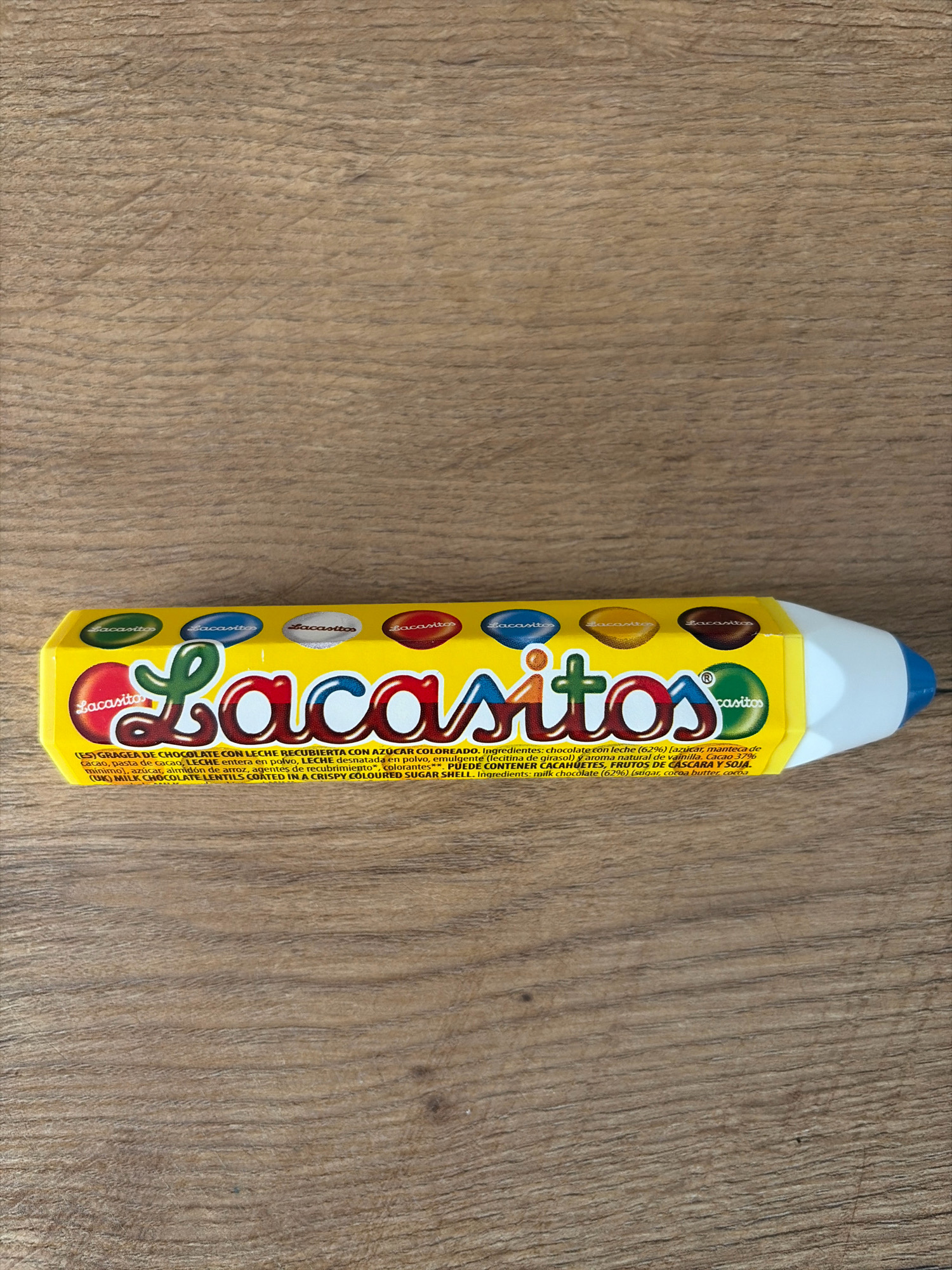 A yellow, pencil-shaped box of Lacasitos candy lies on a wooden surface. The box has a blue tip and white eraser at one end. The front of the box features the brand name "Lacasitos" in colorful lettering and a list of ingredients in both Spanish and English. Several individual Lacasitos candies, each with the brand name, are visible in a line above the main logo. The wooden surface has a prominent grain pattern. The scene is indoors, and there is no indication of the city of Oxentea, Moldova. There are no people or actions depicted in the image.