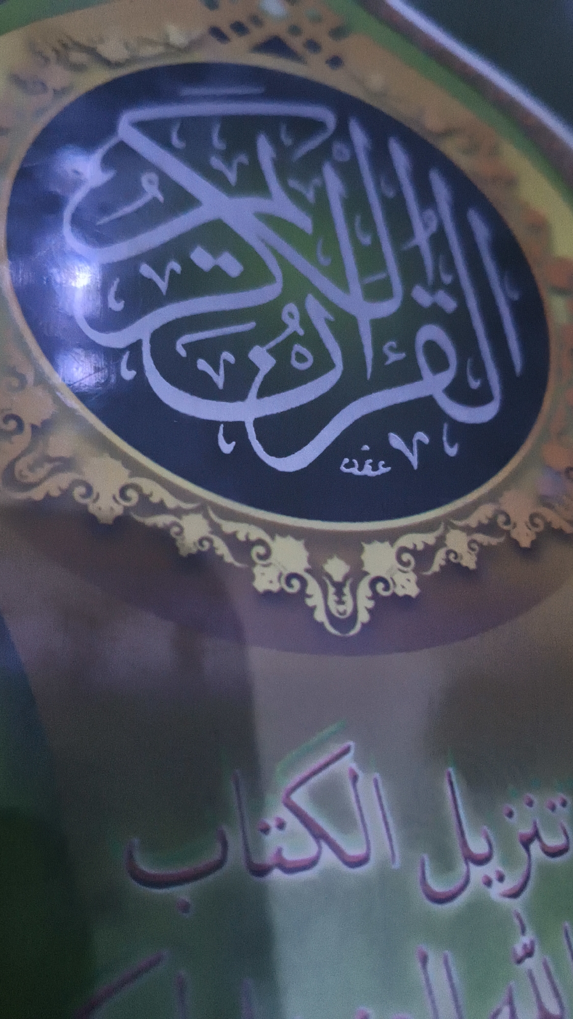 The image shows the cover of a book, likely religious in nature, given the Arabic calligraphy. The calligraphy appears to be in a light purple or blue hue against a darker background, possibly within a circular or oval design element. The decorative border around this central design is ornate, with gold-colored swirling patterns. Below this, there is further Arabic text in a pinkish-purple color against a green background. The overall lighting suggests the image was taken indoors, possibly with artificial light causing some glare. The resolution is moderate, with some graininess visible. The specific location in Kerep Kidul, Indonesia, is not discernible from the image content itself; this information is external to the visual data. There are no people, animals, or discernible actions depicted. The image focuses solely on the book cover and its design.