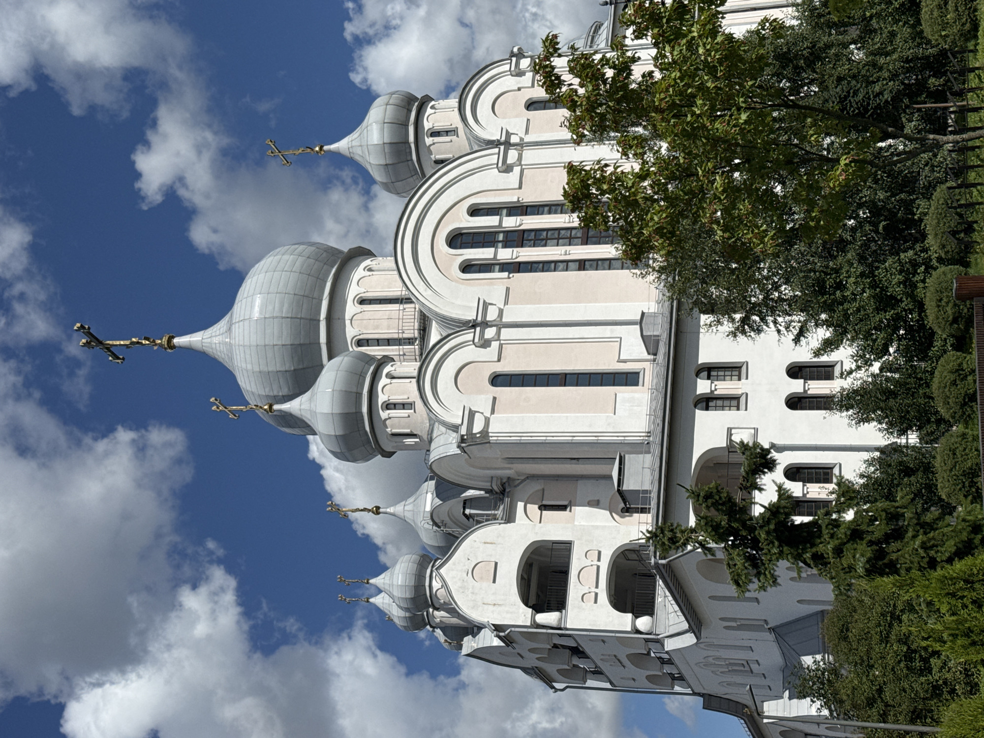 The image shows a view of the Church of the Icon of the Mother of God Joy of All Who Sorrow in Saint Petersburg, Russia. The church is a large, white building with multiple silver domes topped with gold crosses. It has a complex architectural design with several levels, arched windows, and balconies. Lush green trees and foliage surround the base of the church, and a blue sky with white clouds forms the background. The photograph is taken from a low angle, emphasizing the height and grandeur of the church.