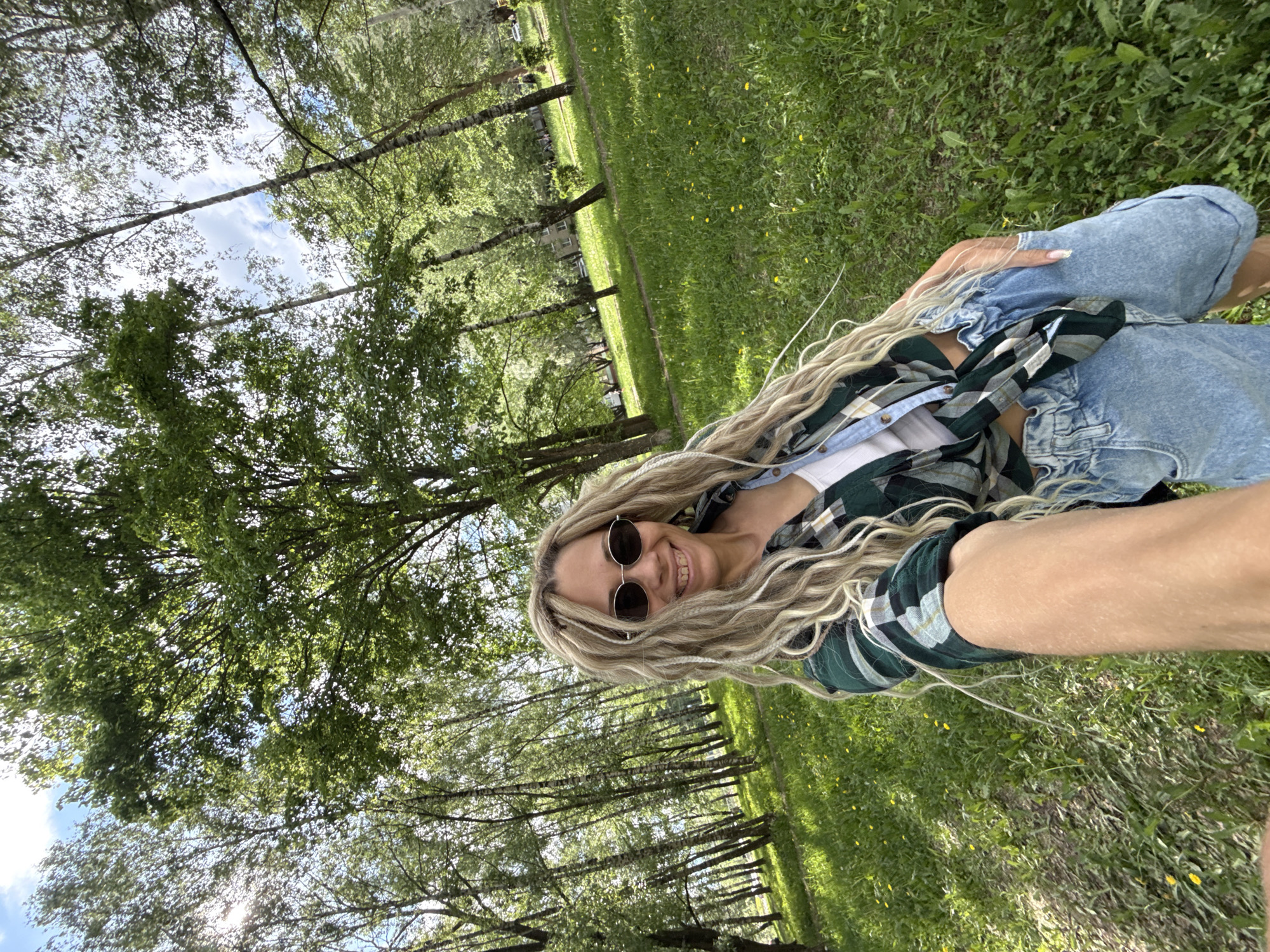 A woman with blonde, wavy hair wearing sunglasses, a green plaid shirt over a white top, and denim shorts smiles for a selfie. The background features a grassy area with trees, possibly a park or wooded area, suggesting an outdoor setting. The photo is taken at a low angle, tilting slightly to the side. The location is Rusanovo, Russia.
