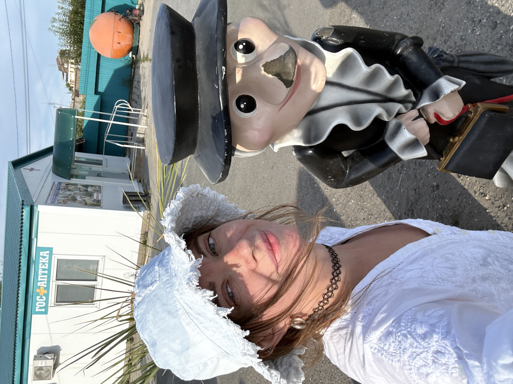 A woman wearing a white embroidered shirt and a light blue bucket hat stands for a selfie next to a garden gnome. The gnome is dressed in a black suit with a ruffled collar and holds a small black briefcase. Behind them is a white building with a green roof, a sign reading "ГОС. АПТЕКА" (State Pharmacy), and a bright orange, orb-shaped object. The location is identified as Sol-Iletsk, Russia. The scene appears to be outdoors, under a clear sky, with some vegetation and a teal-colored fence in the background.