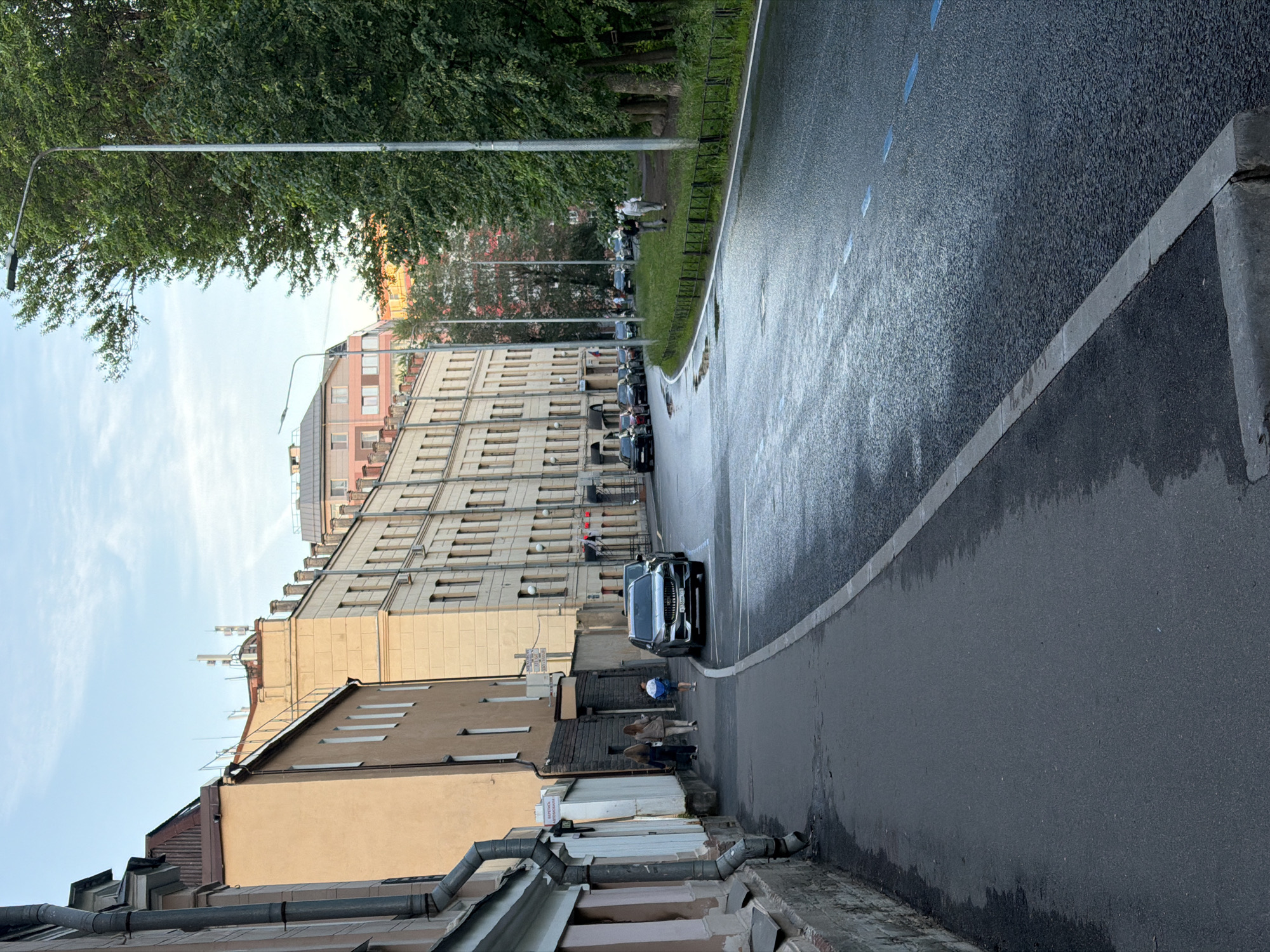 The image depicts a street scene in Saint Petersburg, Russia, on a partly cloudy day. The perspective is from a sidewalk looking down a wet asphalt road that curves to the left. To the left of the sidewalk are buildings with facades of light brown and cream colors. One building has prominent pipes running down its side. Several people are walking on the sidewalk, including two women and a child. A silver car is parked on the right side of the road. To the right of the road is a grassy area with a black metal fence, bordered by tall green trees. A streetlamp is visible in the foreground on the right. The road surface shows reflections from the sky and streetlights, indicating it has recently rained or is damp. In the background, more buildings and trees are visible, with a glimpse of a modern building with red elements. The overall atmosphere is urban, with a blend of older architecture and natural elements.