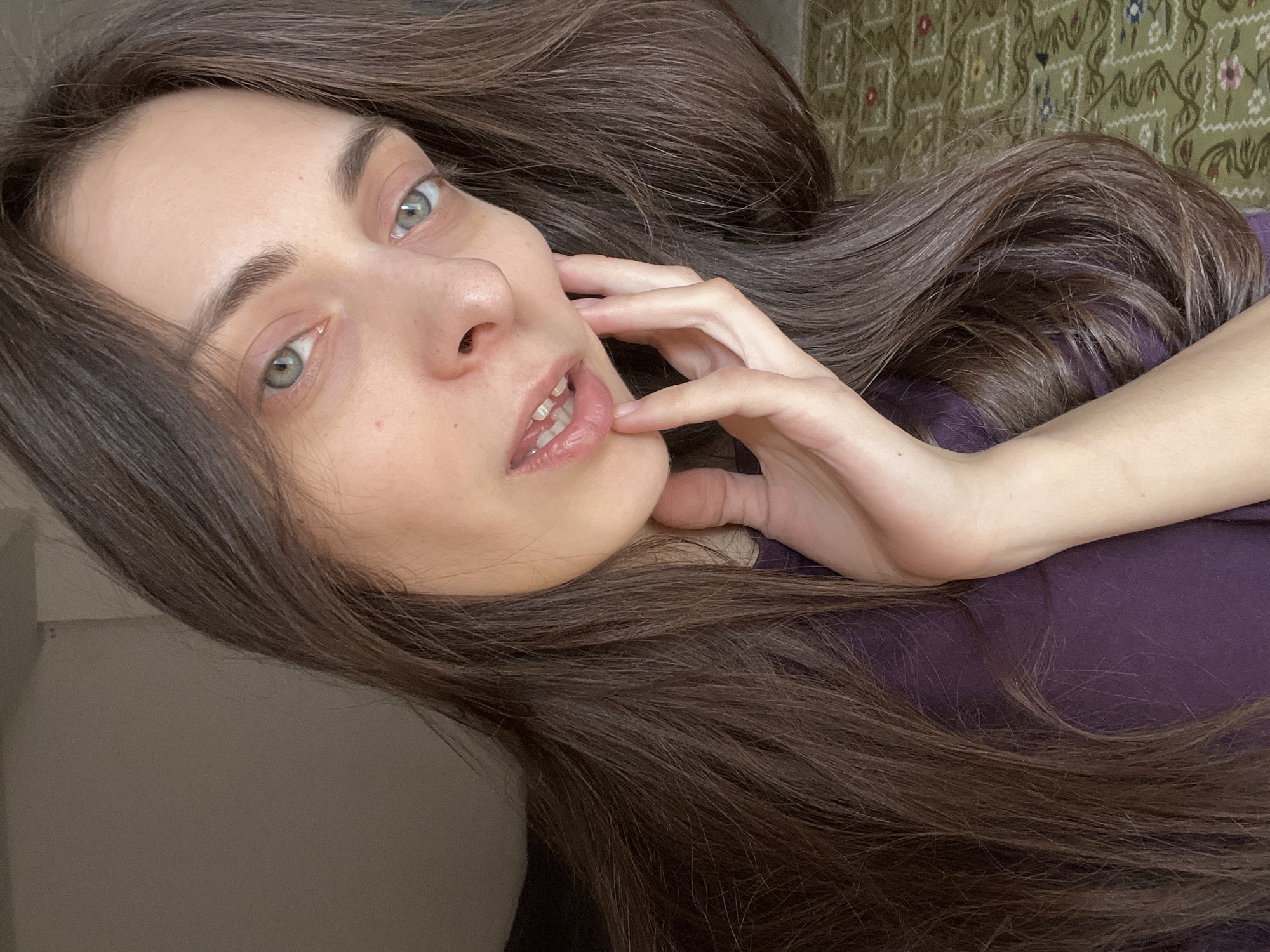 A woman with long, dark brown hair and light-colored eyes is facing the camera, her head tilted slightly. Her hand is near her face, with fingers lightly touching her cheek. She is wearing a dark purple shirt. The background appears to be a wall with a patterned covering or tapestry, and another wall painted white. The image does not provide explicit information indicating the city of Krasnoyarsk, Russia. Therefore, no location details can be inferred.
