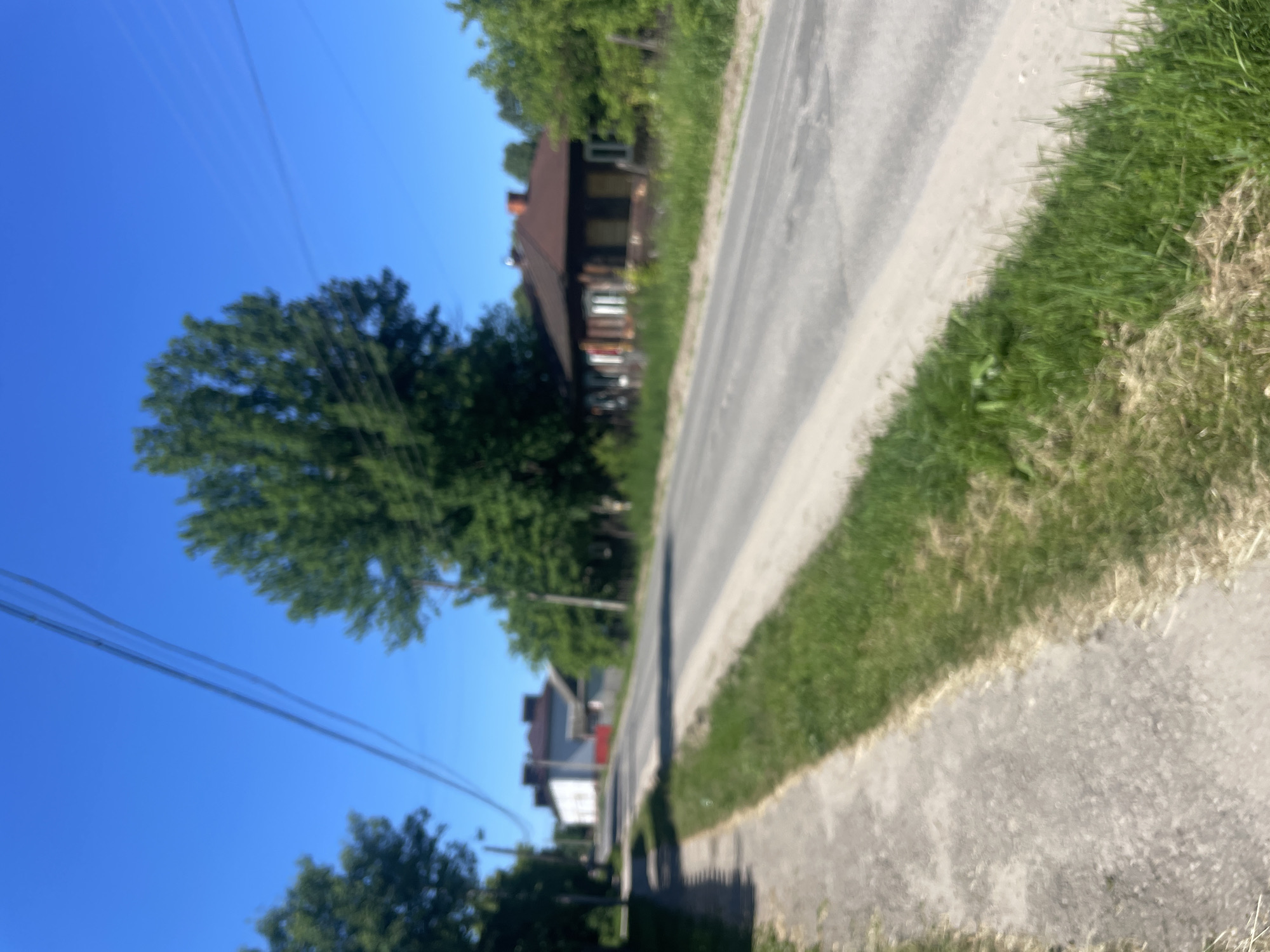 The image shows a street scene in the settlement of Krasnye Baki, Russia, on a clear, sunny day. The street is paved and runs through a residential area. On the left side of the street, there is a concrete sidewalk bordered by grass and trees. On the right side of the street, there are several houses, one of which is a wooden structure with a dark brown exterior and a reddish-brown roof. Another building in the background appears to be larger and white with a dark roof. Overhead, power lines are visible against the bright blue sky. The overall impression is of a quiet, somewhat rural street in a Russian town.