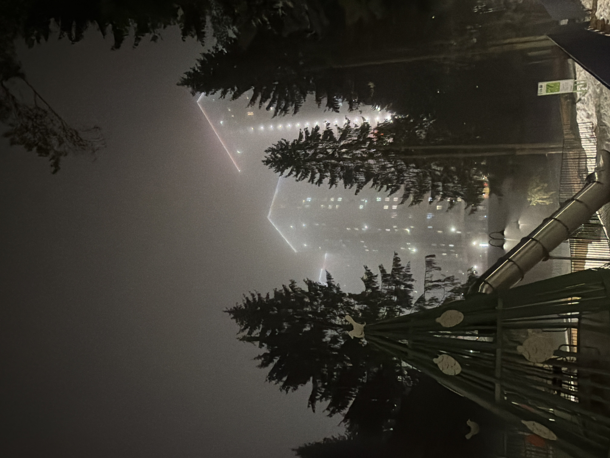 A night scene in Izhevsk, Russia, obscured by heavy fog. Two large buildings with numerous lit windows are visible in the distance, with red laser beams emanating from their roofs. In the foreground, a playground structure, including a metal slide and support frame, is partially visible, with dark silhouettes of trees interspersed. The overall environment appears dark and still, heavily impacted by the fog. A sign with green text is also visible on the right side of the image.