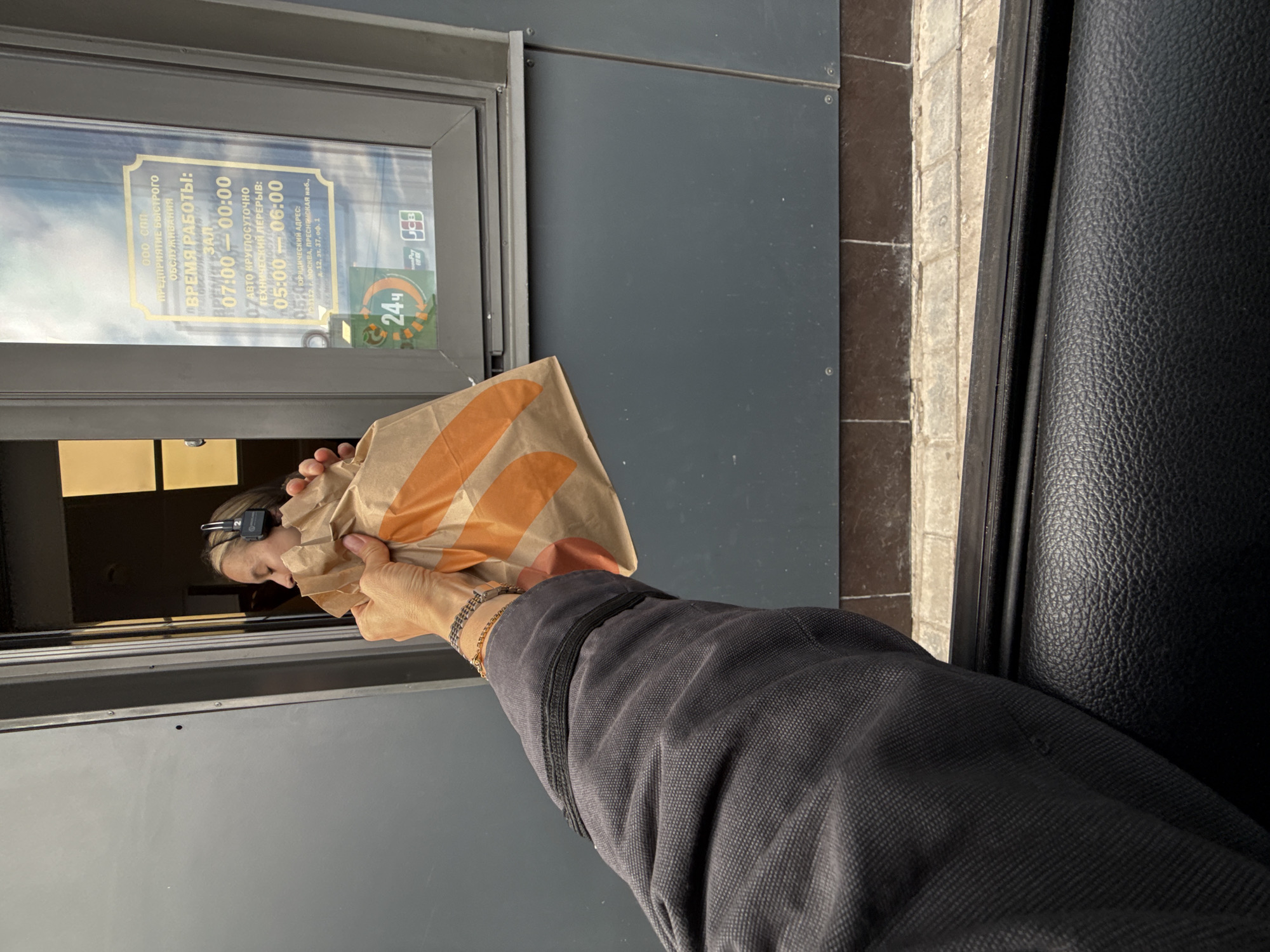 The image shows a hand holding a paper bag with orange accents outside a drive-through window. Inside the window, a person wearing headphones is visible. A sign on the window displays operating hours in Russian, indicating it is a fast-food establishment. The sign reads "OOO CПП ПРЕДПРИЯТИЕ БЫСТРОГО ОБСЛУЖИВАНИЯ" (OOO SPP Enterprise of Fast Service), "ВРЕМЯ РАБОТЫ" (Operating Hours), "ВРЕМЯ ЗАЛ" (Hall Time) 07:00 - 00:00, "АВТО КРУГЛОСУТОЧНО" (Auto 24/7), "ТЕХНИЧЕСКИЙ ПЕРЕРЫВ" (Technical Break) 05:00 - 06:00. Below the operating hours, it lists "ЮРИДИЧЕСКИЙ АДРЕС" (Legal Address) 125110, г. Москва, Пресненская наб., д. 12, эт. 37, оф. 1. There is no specific information in the image that can place the location in the city of Izhevsk, Russia. The scene appears to be taken from inside a vehicle looking out towards the drive-through window.