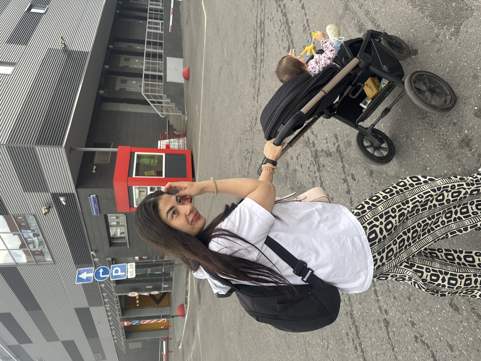 In a Moscow city scene, a woman with long dark hair, wearing a white t-shirt, black backpack, and black and white patterned pants, pushes a black stroller containing a baby. The baby is wearing a pink floral outfit and holding a yellow toy. The background features a modern building with a grey corrugated facade and a red security booth. Street signs, including an upward arrow and a parking sign, are visible on the left. The ground is asphalt, and the overall atmosphere is that of an urban environment.
