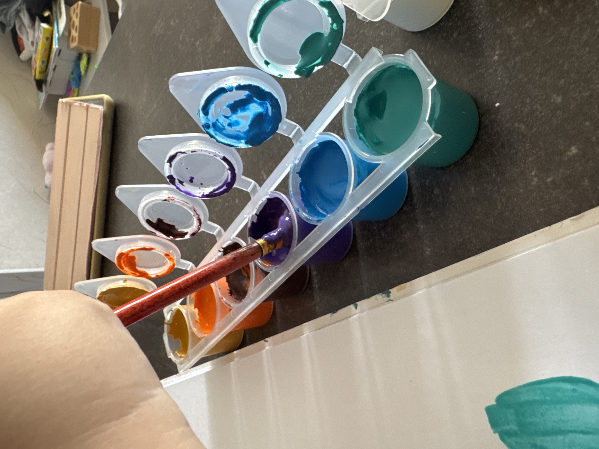 A hand holding a paintbrush is shown dipping the brush into a small container of purple paint. The paint pots are arranged in a white plastic holder, and there are various colors of paint visible, including orange, yellow, brown, purple, blue, and green. A white sheet of paper is positioned to the left of the paint pots, with some green paint already applied to it. The scene appears to be set on a dark, speckled countertop. In the background, there are some objects on shelves, including what looks like a wooden block and some boxes. The overall impression is of someone engaged in painting or a related art activity. The image does not provide any information about the location being the city of Верхнерусское, Russia.