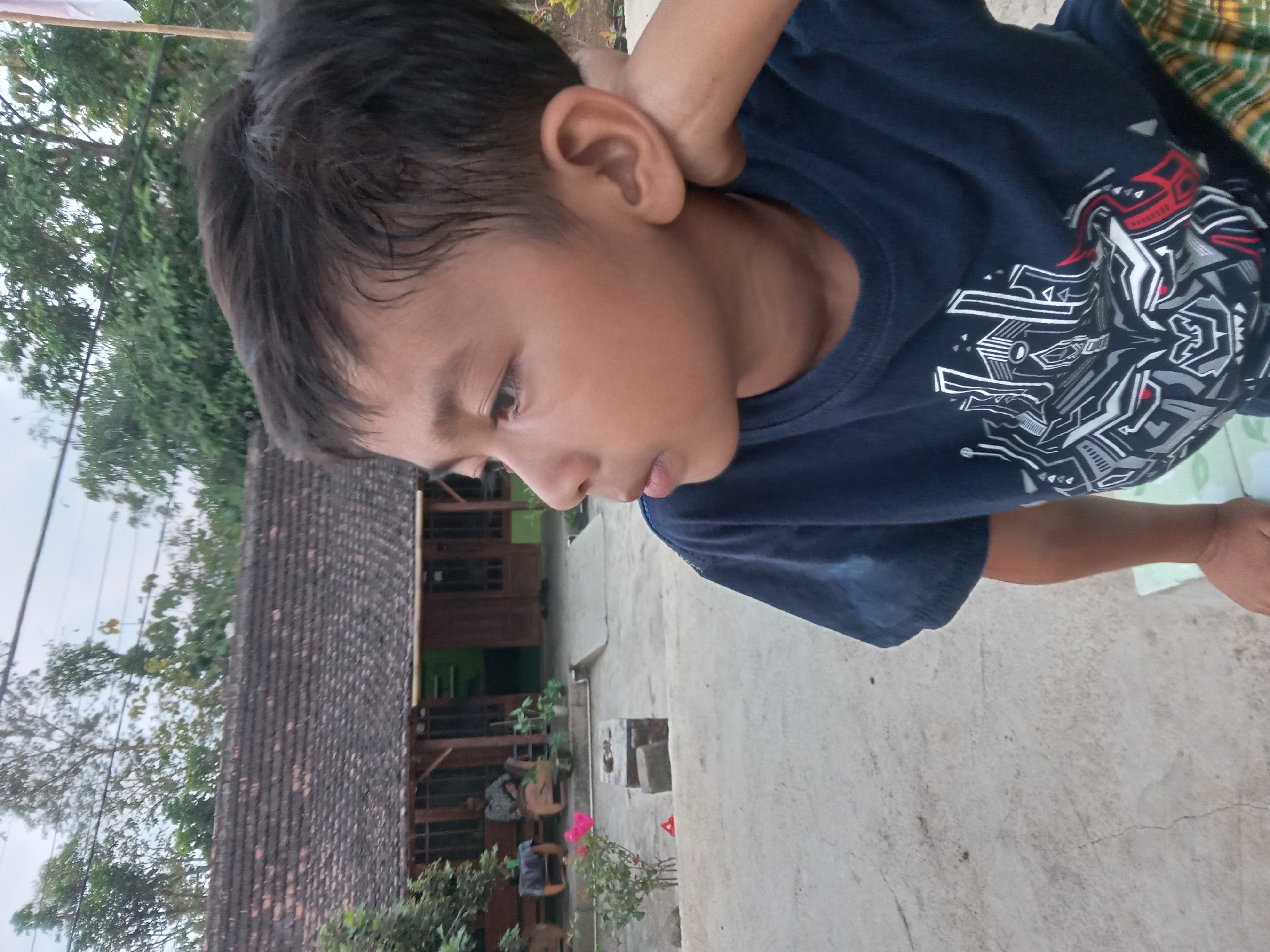 A young boy is photographed in close-up, facing right and looking away from the camera. He has short, dark hair and is wearing a dark blue t-shirt with a graphic design on it. His right hand is resting on the back of his head. The background suggests a rural or semi-rural setting in Kedunggudel, Indonesia, with a traditional house with a tiled roof visible. Trees and vegetation are also present. The ground appears to be concrete or a similar paved surface.