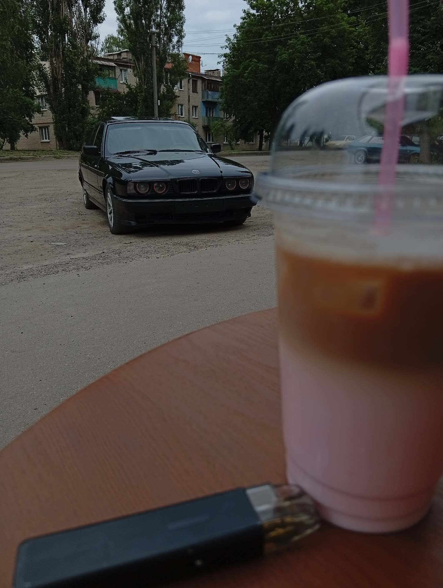 In an outdoor urban setting, a black BMW E34 sedan is parked on a dirt lot beside a paved road. In the foreground, on a wooden table, is a clear plastic cup with a pink lid and straw, containing a layered beverage, and a black electronic cigarette. Behind the car, a building with multiple stories and windows is visible, surrounded by trees. The sky is overcast.