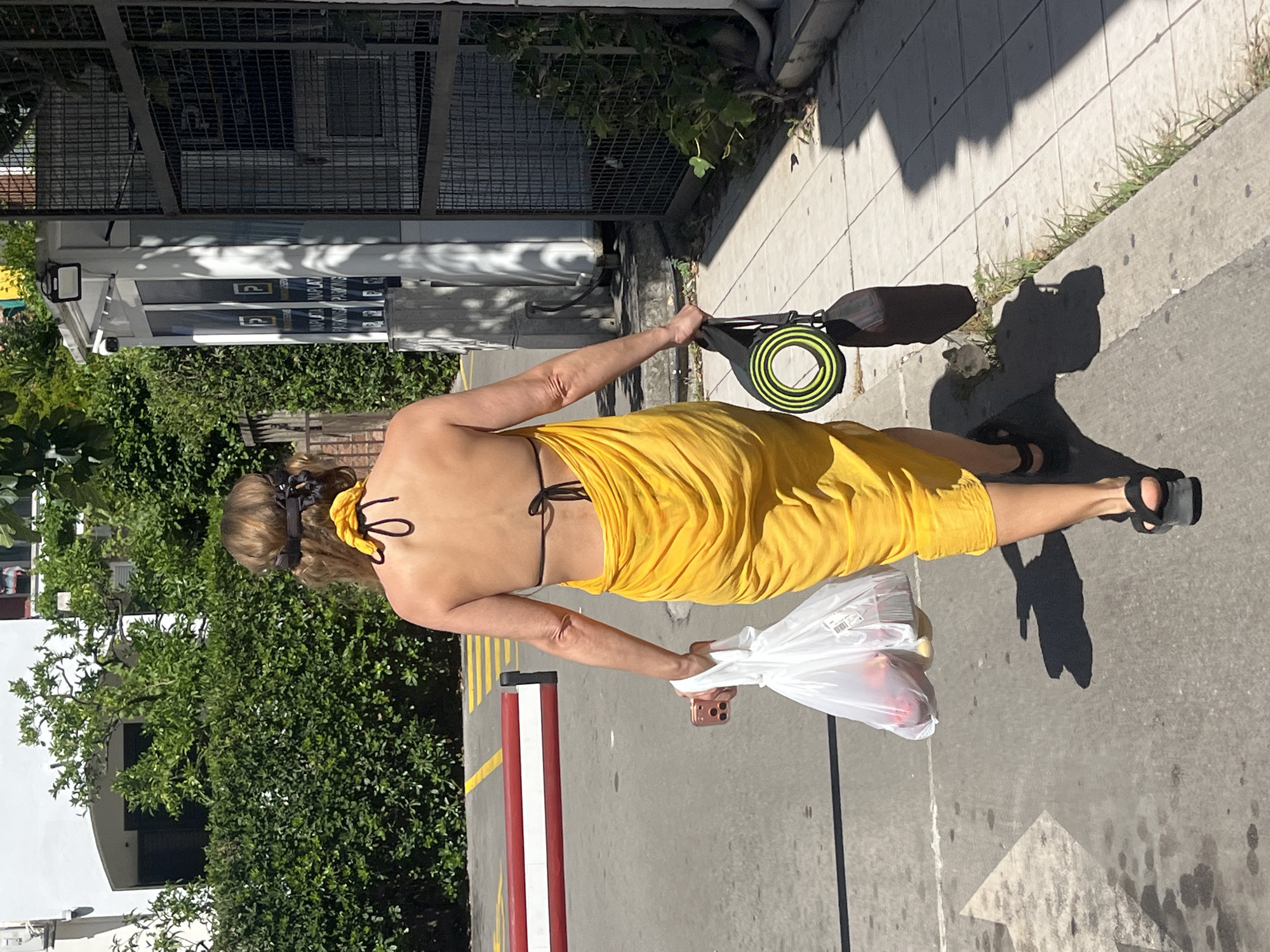 A person wearing a yellow sarong and a black bikini top walks down a sidewalk. They are carrying a white plastic bag filled with groceries in their right hand and a coiled yoga mat in their left. The person has brown hair pulled back with a black and yellow accessory. Sunlight casts long shadows on the concrete, indicating it is likely daytime. Green foliage and a building with a metal fence are visible in the background. A white and red pole is also present on the sidewalk. The scene appears to be a casual outing in a public space.