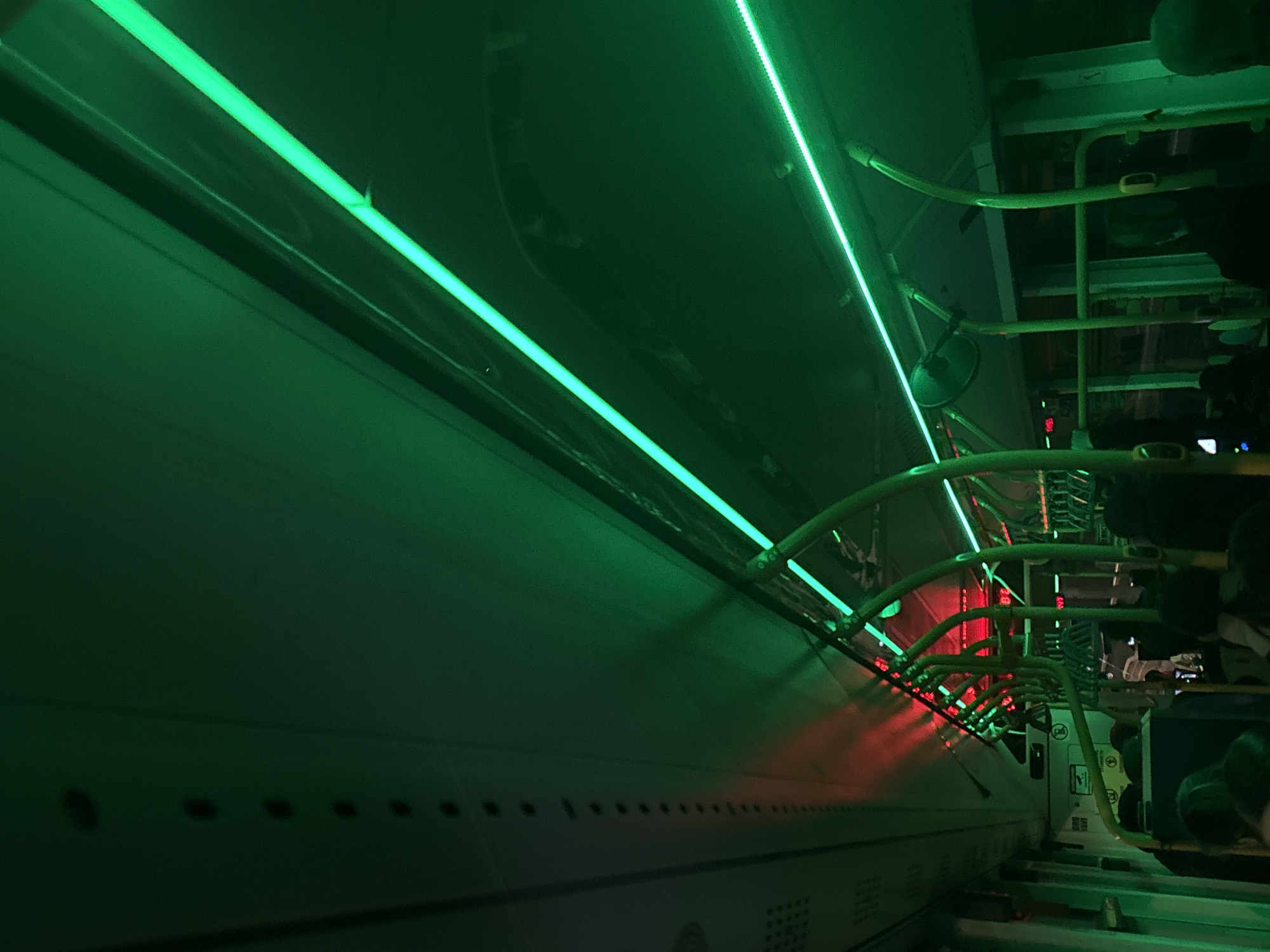The image depicts the interior of a submarine-themed attraction in Varna, Bulgaria. A curved wall of the submarine extends along the left side of the frame, illuminated by green neon lights. A series of small, circular openings is visible along the lower part of the wall. Overhead, a network of yellow metal pipes and handrails runs parallel to the wall, also illuminated by green neon lights. Towards the right side of the frame, figures are visible in the dim light, possibly visitors or staff. A red glow is visible in the distance, suggesting another lighting element.