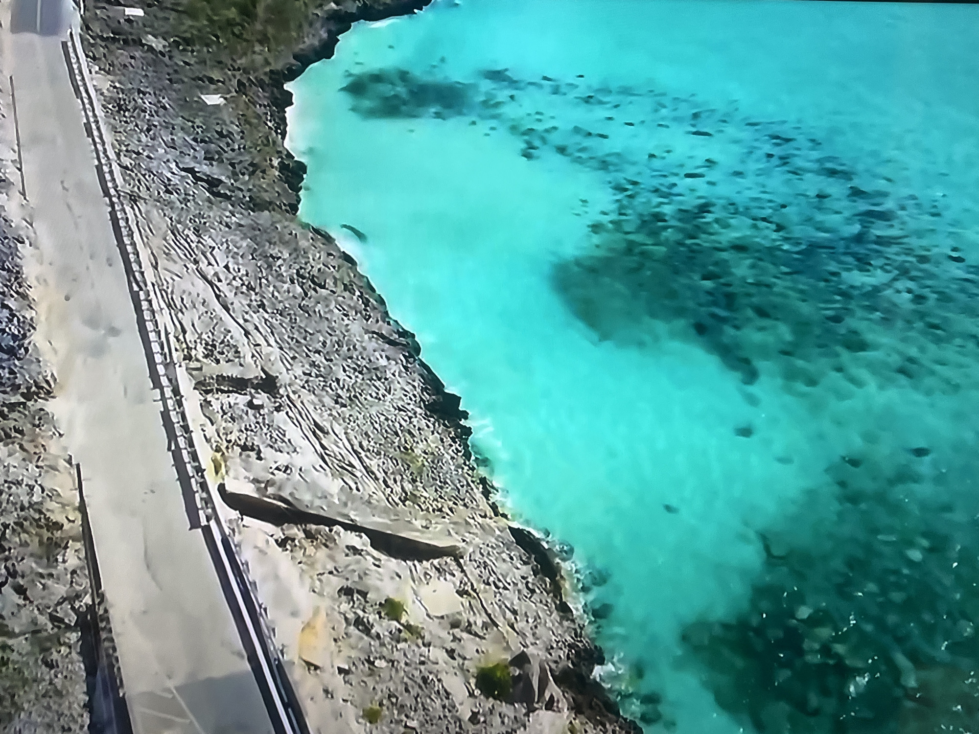 An aerial, high-angle shot shows a paved road running alongside a rocky coastline and turquoise water. The road is gray and appears to have a guardrail or fence along its edge closer to the water. The coastline is composed of dark, jagged rocks with patches of green vegetation. The water is a vibrant, clear turquoise and appears to have shallow areas with visible coral or seabed features. The scene suggests a remote, coastal environment, possibly a scenic overlook or a road along a tropical island.