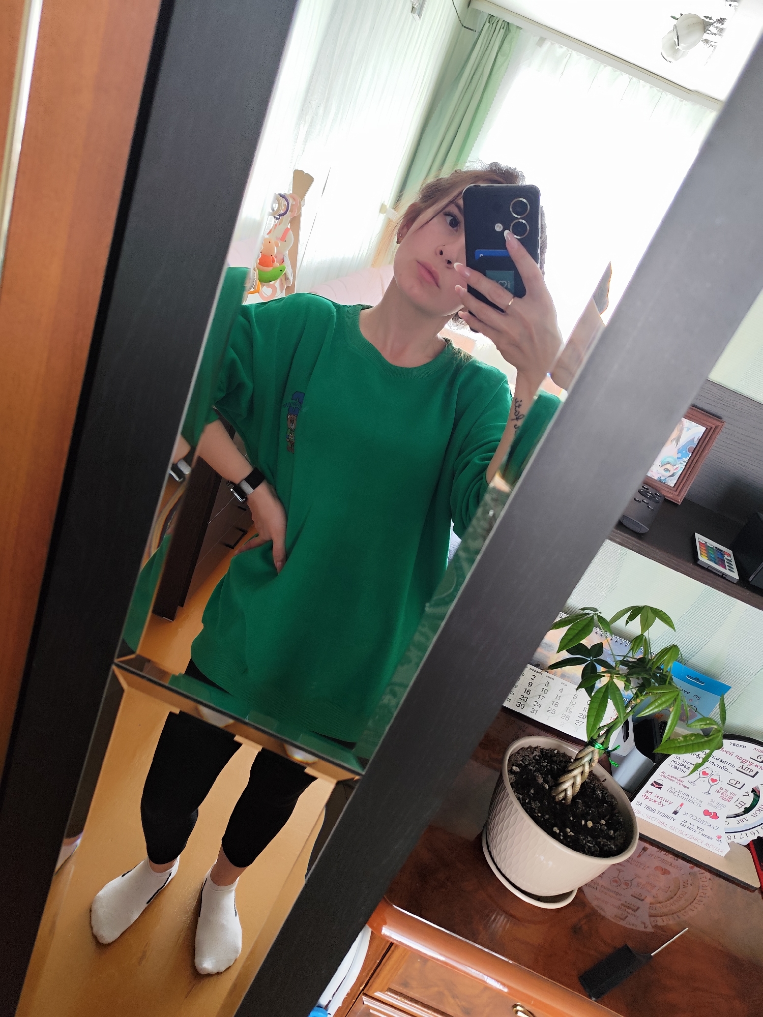 The image is a mirror selfie of a woman indoors. She is holding a phone in her right hand, obscuring part of her face. She has fair skin, light brown hair, and is wearing a green oversized sweatshirt, black leggings, and white socks. She's standing in front of a mirror that reflects the room around her. The room appears to be a bedroom with a window in the background. Near the mirror, there is a table with a potted plant, a calendar with some text in Russian ("ТВОРИ", etc.) visible, and a framed picture on a shelf. The location is indicated in the OCR as Фоки, Russia. The mirror frame is dark, and the wooden furniture is a mix of brown and tan tones.