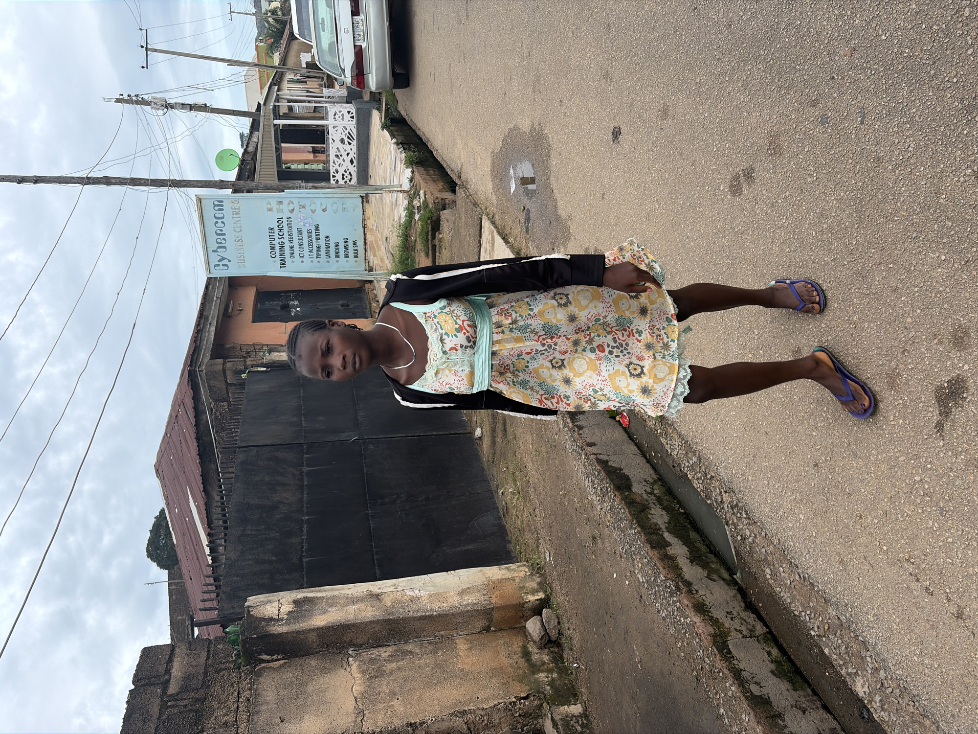 A young Black female walks on a paved street in a residential or commercial area in Jos, Nigeria. She is wearing a floral print dress with a light blue sash, a black jacket, and purple flip-flops. The street is lined with a concrete drainage ditch on the left and buildings on the right. A sign for "Cybercom Business Centre & Computer Training School" is visible in the background. The sky is overcast. A silver car is parked on the right side of the street.