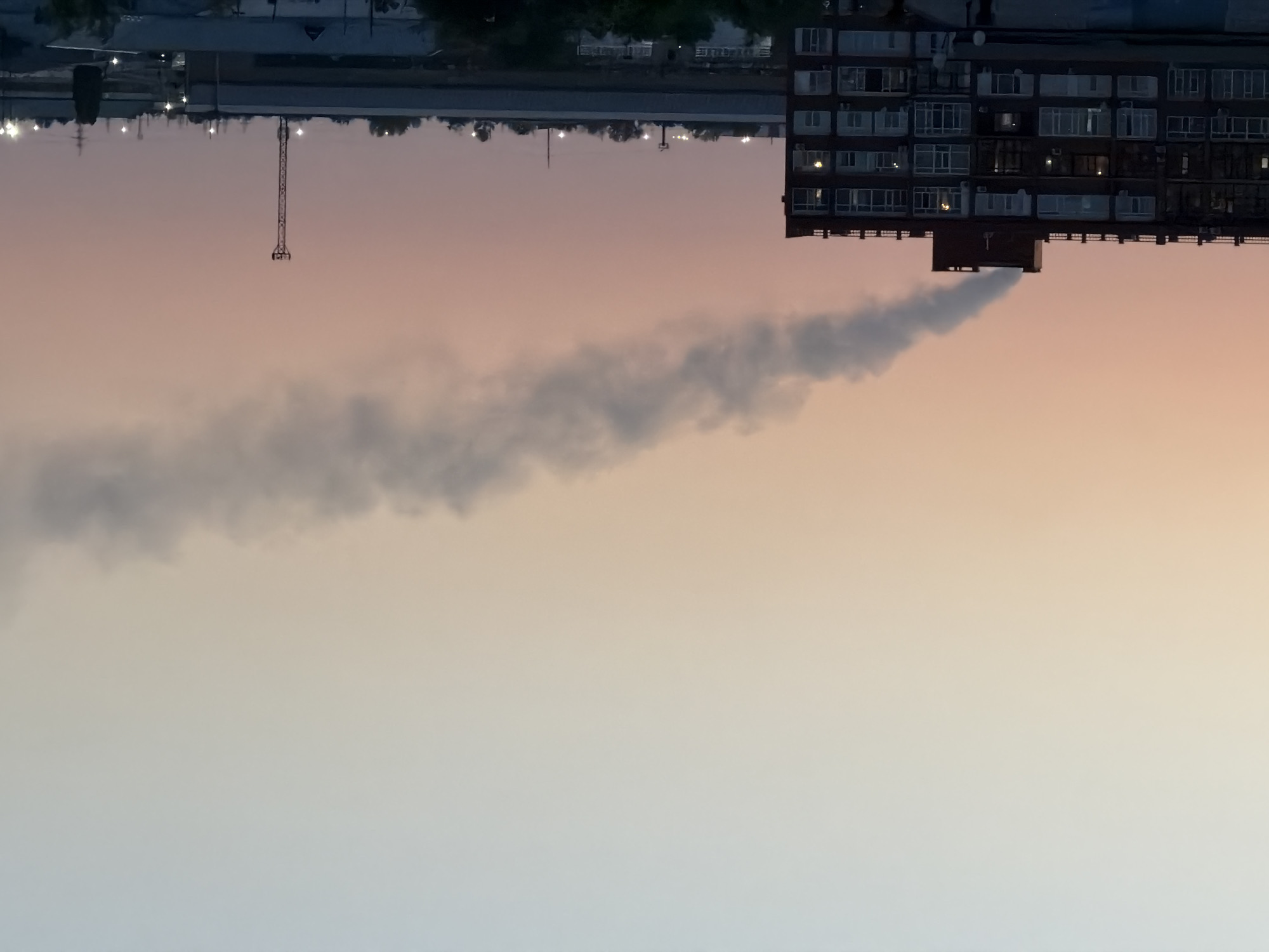 The image shows a building with smoke emanating from its roof against a twilight sky. The building is a multi-story apartment complex with numerous windows. A tall, slender transmission tower is visible in the distance, along with other industrial or commercial structures. The sky is a gradient of pale blue and pink, suggesting it is either dawn or dusk. The scene appears to be an urban or industrial area. The location is identified as the city of Pavlodar, Kazakhstan.