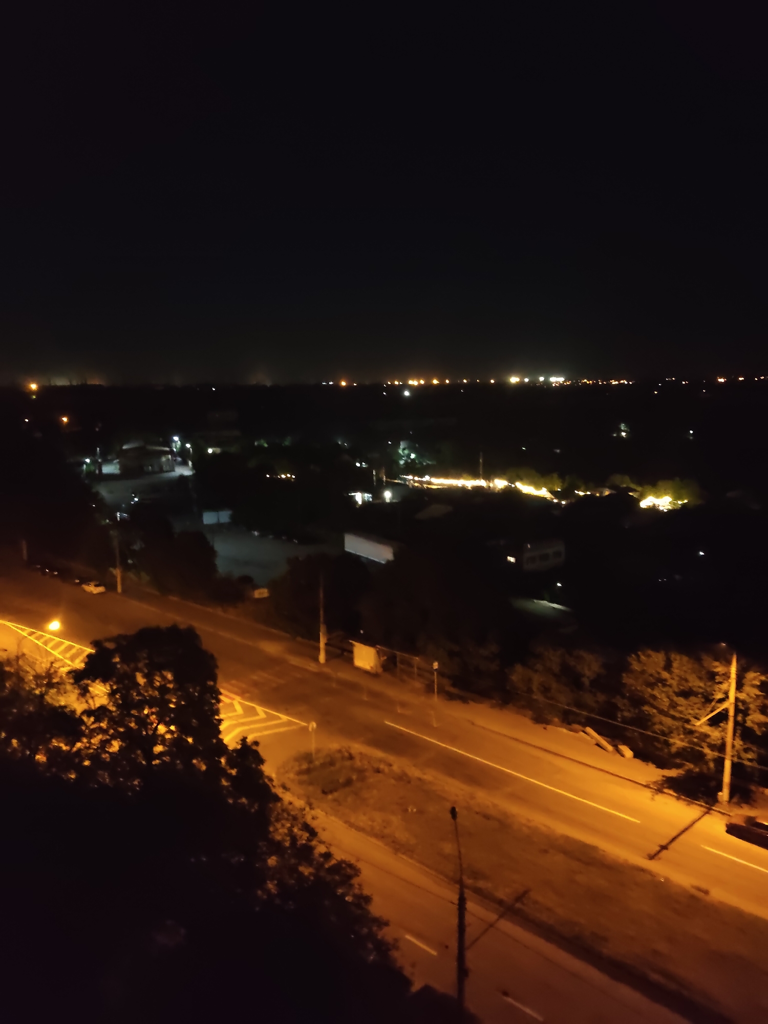 A nighttime aerial view captures a suburban area in Zaporizhzhia, Ukraine, illuminated by artificial lights. A two-lane road runs diagonally through the lower portion of the frame, with bright yellow streetlights casting long shadows and highlighting white road markings. Trees and other vegetation line the road and the surrounding landscape. In the middle ground, several buildings, possibly houses or small industrial structures, are visible, their outlines softened by the darkness and illuminated by sparse lights. In the far distance, a faint glow indicates a more densely populated or industrialized area. The overall scene depicts a quiet, nocturnal urban periphery.