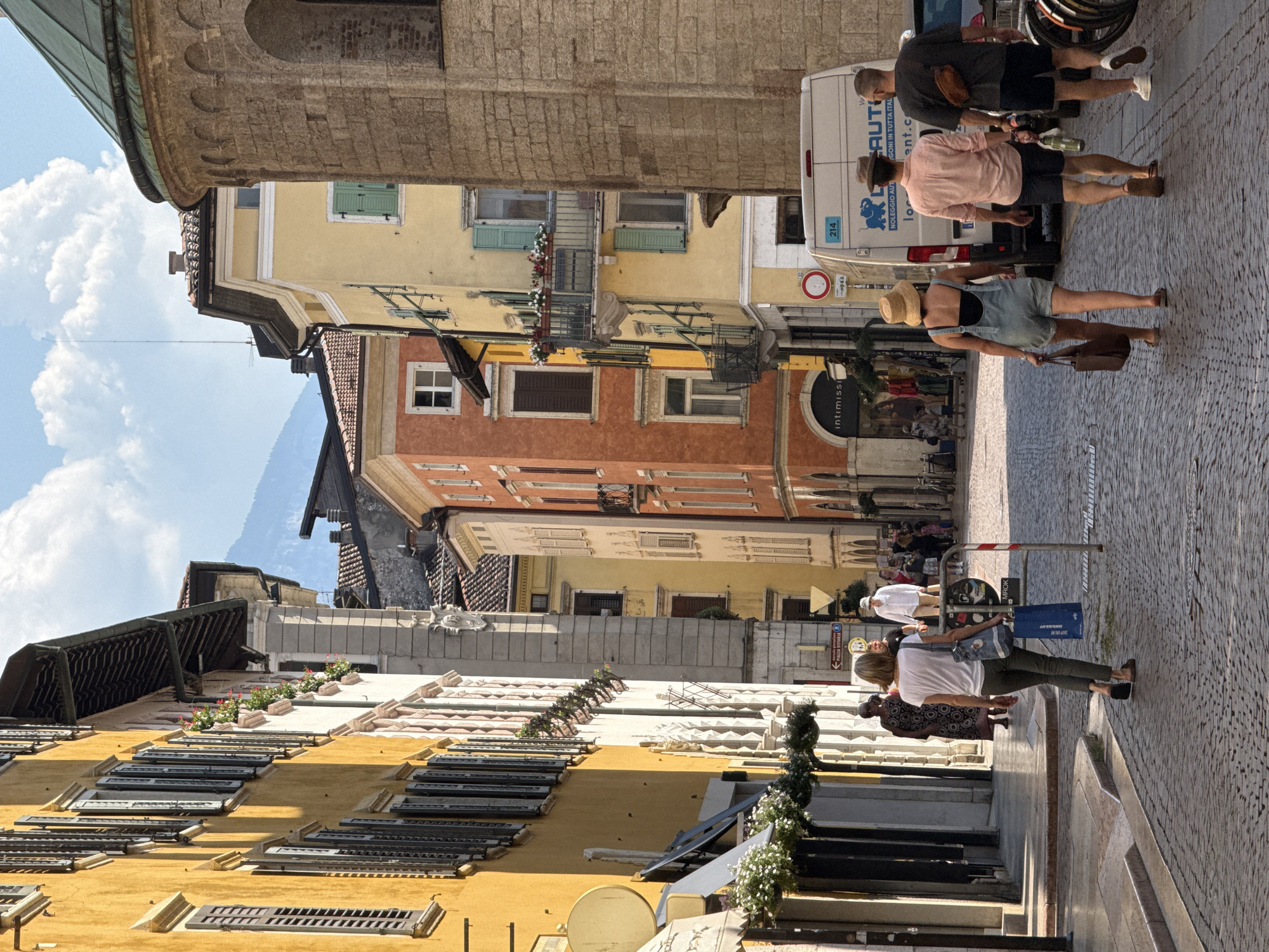 This image captures a sun-drenched street scene in Trento, Italy, likely in the historic city center, with tall, colorful buildings lining a cobblestone street. On the left, a bright yellow building with numerous closed dark shutters dominates the foreground, featuring awnings and hanging plants. Further down the street, buildings display orange, yellow, and cream facades, adorned with balconies and more greenery.
In the mid-ground, a white van with the text "Noleggio Auto" is parked, suggesting a vehicle rental service. Several people are walking along the street. Closest to the viewer, a woman in denim overalls and a straw hat walks away from the camera, carrying a brown bag. Behind her, two men are also walking away. One man wears a pink button-down shirt and dark shorts, while the other wears a dark t-shirt and shorts, with a brown satchel slung across his back. Another woman in a white t-shirt and green pants walks away from the camera in the middle left of the image, carrying a blue shopping bag. Further down the street, other individuals and shoppers are visible, along with bicycles. The street is paved with cobblestones, and the architecture suggests an old European town. A large stone tower with a green dome, possibly part of a church or historical building, is visible on the right side of the image. The sky is partly cloudy and a mountain range is faintly visible in the background, indicating the city's proximity to the Alps.