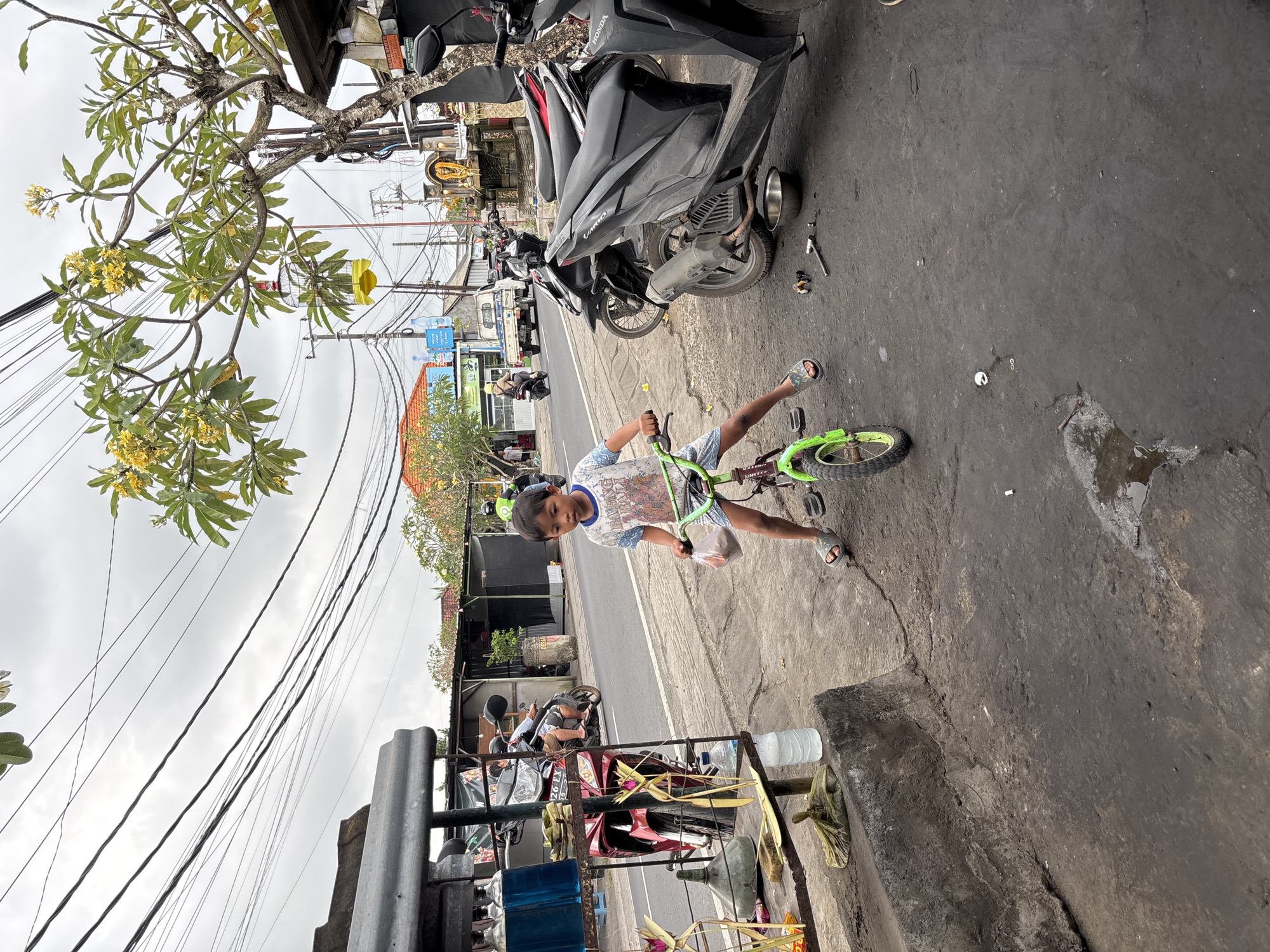 A young boy rides a green bicycle on a paved road in Kuta Selatan, Indonesia. He is wearing a white t-shirt with a graphic print and blue shorts, and gray sandals. He is holding a plastic bag in his right hand. The road is lined with shops and parked motorcycles, with power lines overhead. The sky is overcast. The boy is the main subject of the image, and the environment is a typical street scene in a Southeast Asian city.