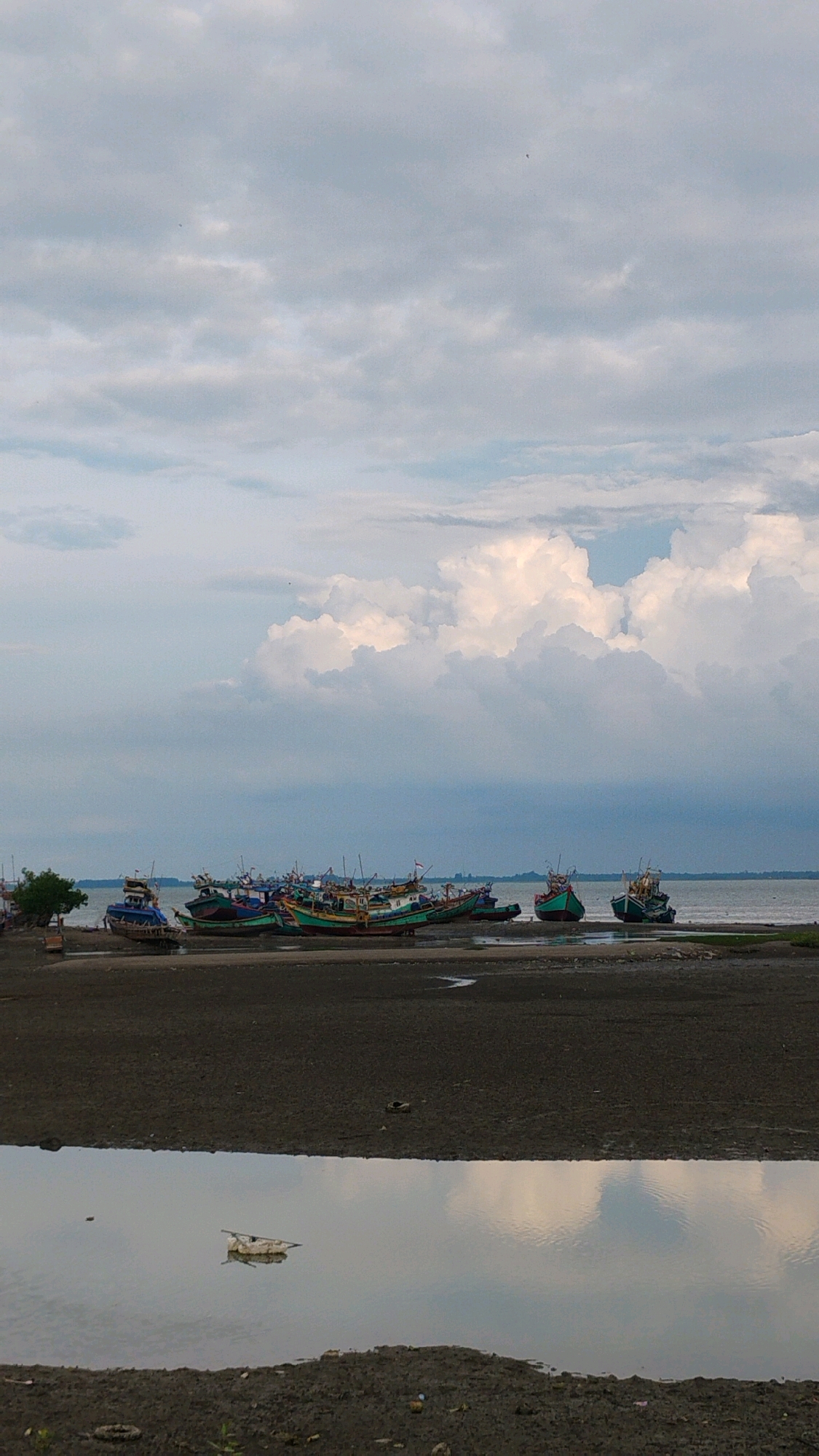 A serene seascape captures a fleet of fishing boats docked along the shore of Lhokseumawe, Indonesia. The sky is a canvas of soft, billowing clouds, with a few small birds dotting the expanse. The boats, painted in various shades of green, blue, and red, rest on the sandy, wet shore, some partially submerged in shallow water. The calm sea stretches out to the horizon, meeting the pale blue sky. In the foreground, a small pool of water reflects the sky and clouds, with a piece of debris floating in it. The overall atmosphere is peaceful and tranquil, typical of a quiet morning or late afternoon in a coastal Indonesian town.