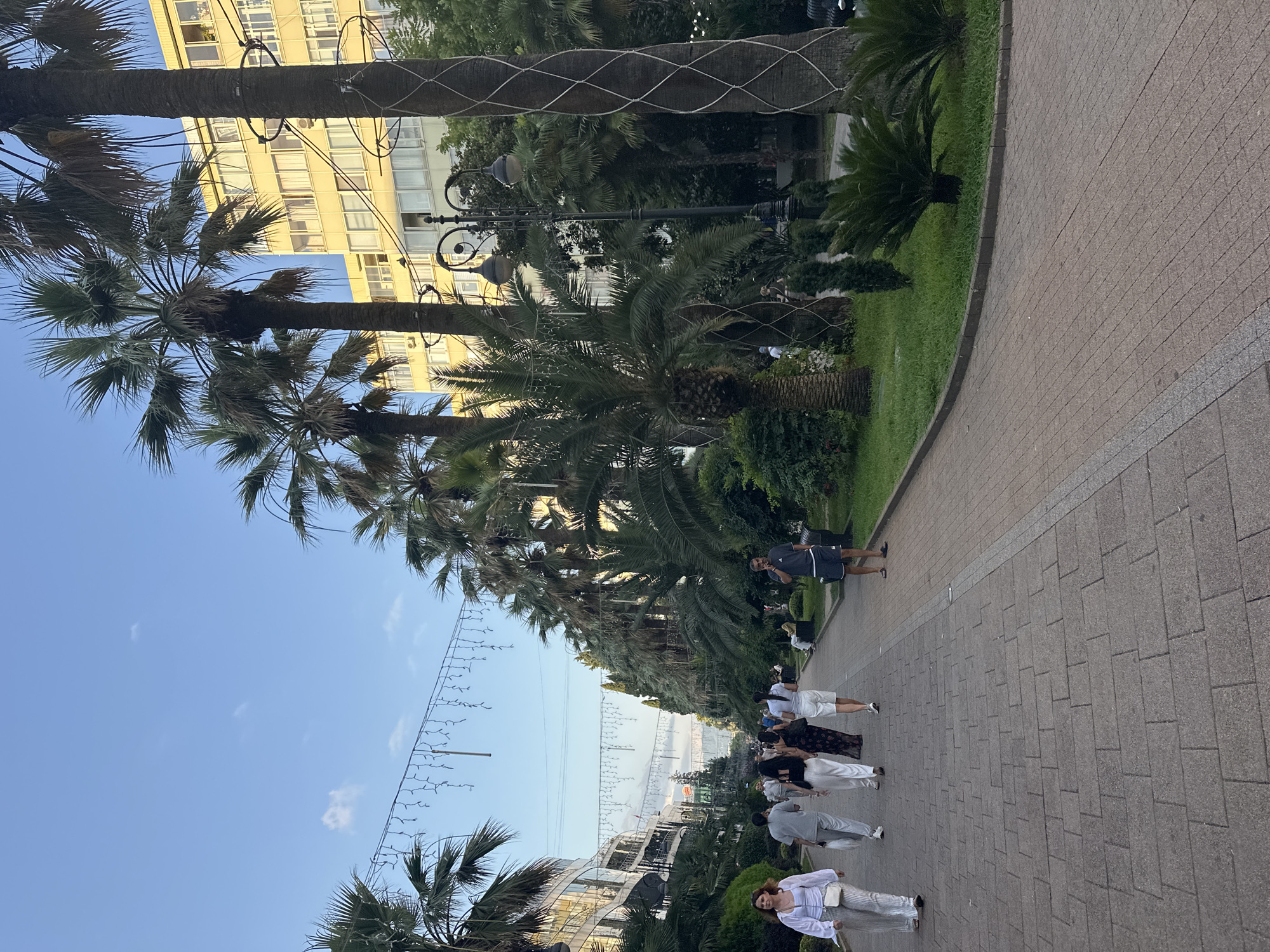 The image depicts a sunlit promenade in Sochi, Russia, lined with lush palm trees and green foliage. A pedestrian walkway, paved with gray and pinkish-brown bricks, curves into the distance. Several people are strolling along the path. On the left, a woman in a white top and striped pants walks towards the viewer, her white crossbody bag visible. Behind her, a group of people are walking away from the camera, including a man in a gray t-shirt and light pants, a woman in a black dress, and a woman in a white t-shirt and white shorts. On the right side of the promenade, a man in a dark t-shirt and shorts stands looking at his phone. In the background, a tall, yellow building with many windows stands prominently, its architecture suggesting it is an apartment building or hotel. String lights are suspended across the promenade, hinting at evening festivities or decoration. The sky is a clear blue with a few wispy clouds. The overall scene conveys a relaxed, leisurely atmosphere typical of a seaside resort town.
