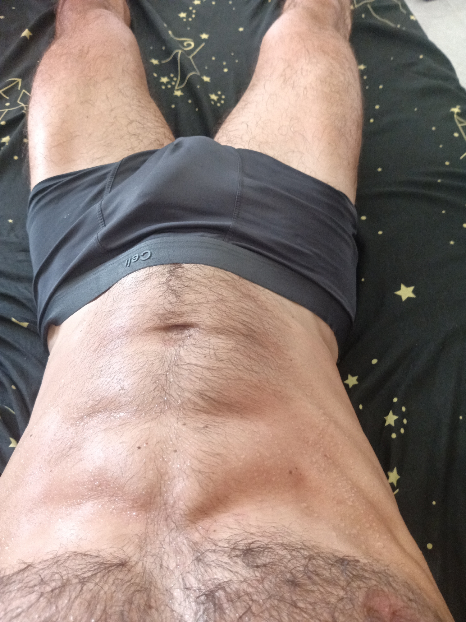 The image shows a close-up, overhead view of a person lying on a bed with a dark, star-patterned comforter. The person is wearing dark gray boxer briefs. Their torso and upper thighs are visible, with visible sweat on the skin. The person's abdomen displays a moderate amount of dark hair, and their thighs are also hairy. The scene is a private indoor setting, and there is no indication of the city of Mũi Né, Vietnam, or any other specific location.