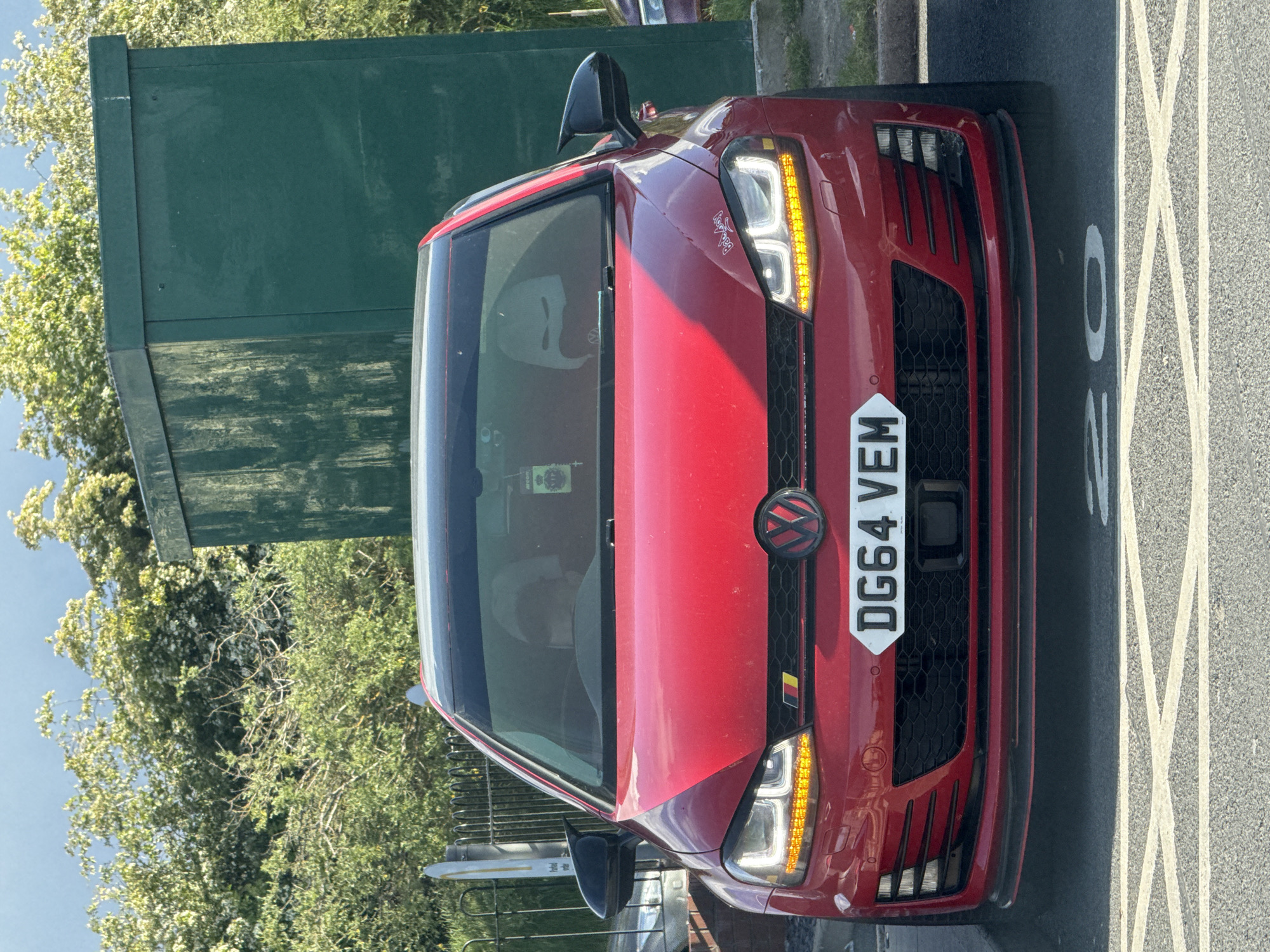 A red Volkswagen Golf with registration number DG64 VEM is parked next to a green electrical box on the roadside in Great Yarmouth, United Kingdom. The front headlights are illuminated. A bicycle rack is visible in the background. The vehicle is parked on a marked road surface with the number "30" painted on it. Trees and foliage surround the scene.