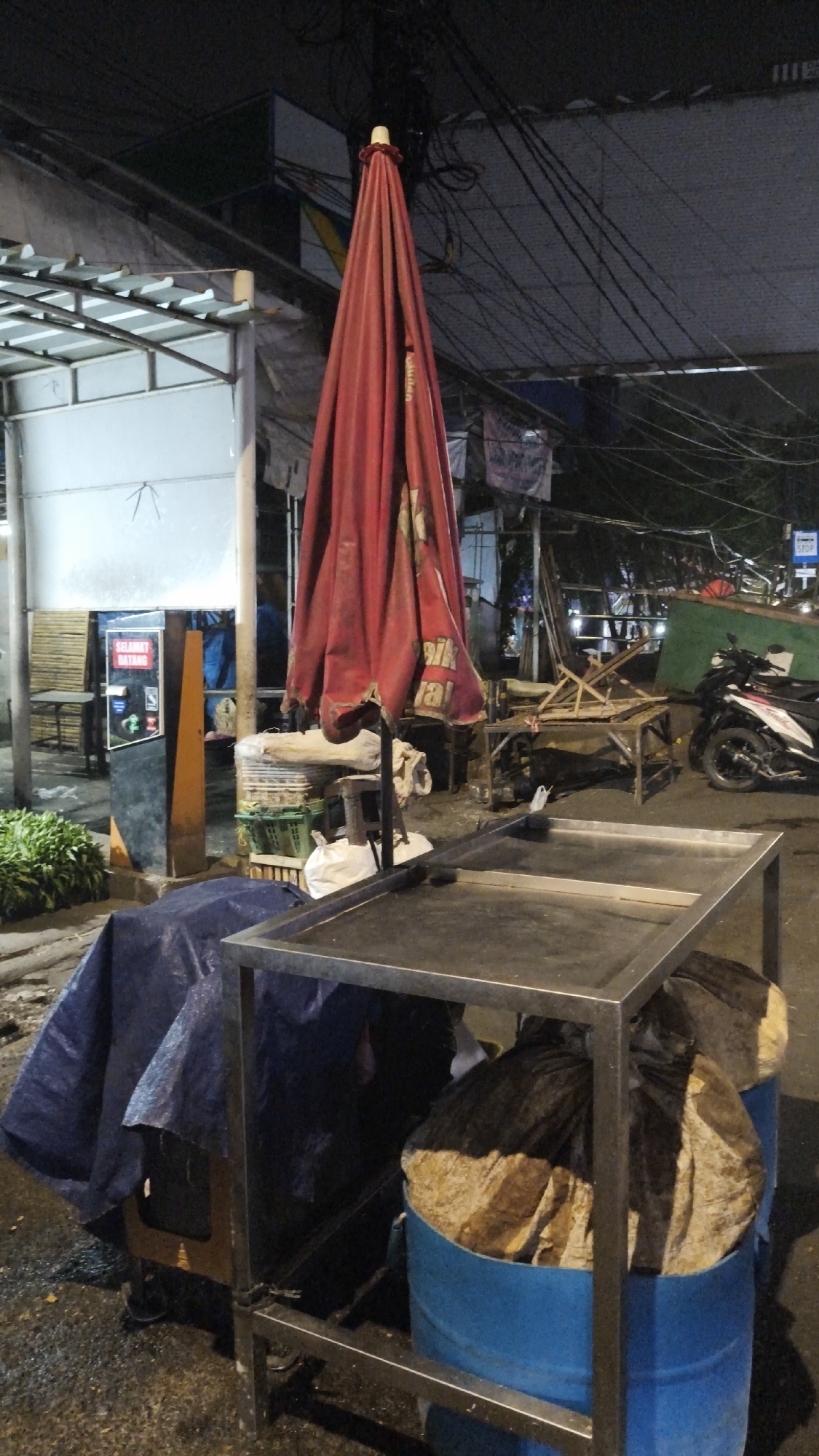 A red umbrella, closed and tilted, dominates the center of the frame, its fabric bunched and creased. It is supported by a dark metal pole. In the foreground, a stainless steel cart with two levels is positioned. The top surface is bare, while the lower level is obscured by items covered in dark plastic bags and blue barrels, some of which contain large, light-colored objects. To the left of the cart, a large blue tarp covers a portion of an unseen object or structure. Behind the umbrella, a variety of items are visible: stacked crates, white plastic bags, and wooden structures. A sign with the Indonesian text "SELAMAT DATANG" (Welcome) is mounted on an orange and black panel to the left. A motorcycle is parked on the right side of the image. The scene appears to be an outdoor marketplace or street vendor area at night, indicated by the artificial lighting and overhead wires. The ground is wet, suggesting recent rain. The overall impression is one of a temporary setup, possibly closed for the night, in an urban environment. The location is identified as South Jakarta, Indonesia.