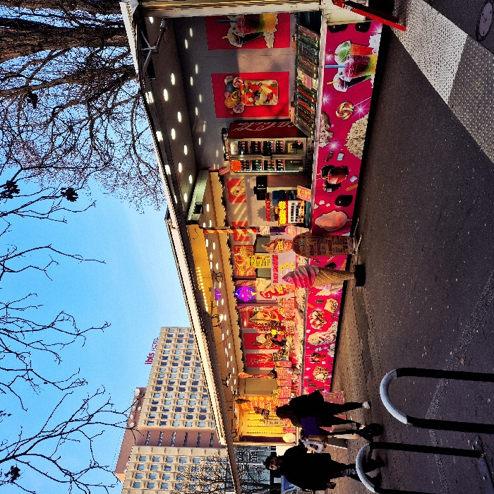 A street scene features a brightly lit food stall or stand adorned with colorful images of ice cream, candy, and drinks. Two people walk past in the foreground. In the background, a tall building with a red "Ibis" sign is visible. The left side of the frame is filled with bare tree branches against a blue sky. The stall appears to be parked alongside a street or sidewalk, with a glimpse of asphalt on the right.