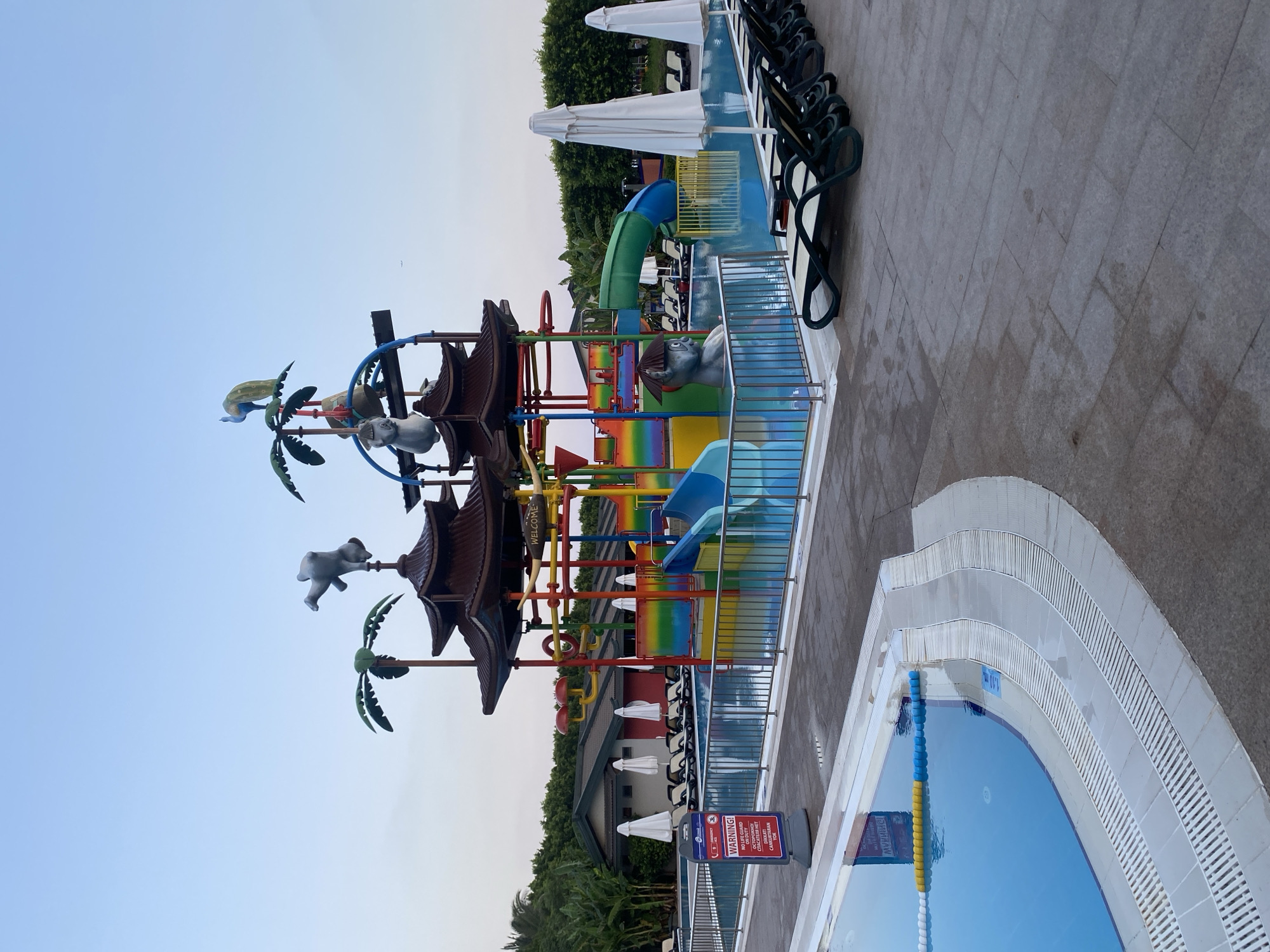 This is a daytime, eye-level, full shot of a water park area in Aksu, Turkey. The foreground features a portion of a bright blue swimming pool with a white and yellow floating rope. To the right of the pool is a tiled walkway with rows of empty dark green lounge chairs and closed white umbrellas. Further in the background is a large, colorful water play structure with slides, palm tree decorations, and animal figures. A red and white "WARNING!" sign stands near the pool. Behind the water structure, there are some buildings with red accents and dark roofs, surrounded by lush green trees. The sky is clear and pale blue, with a faint airplane visible in the distance.