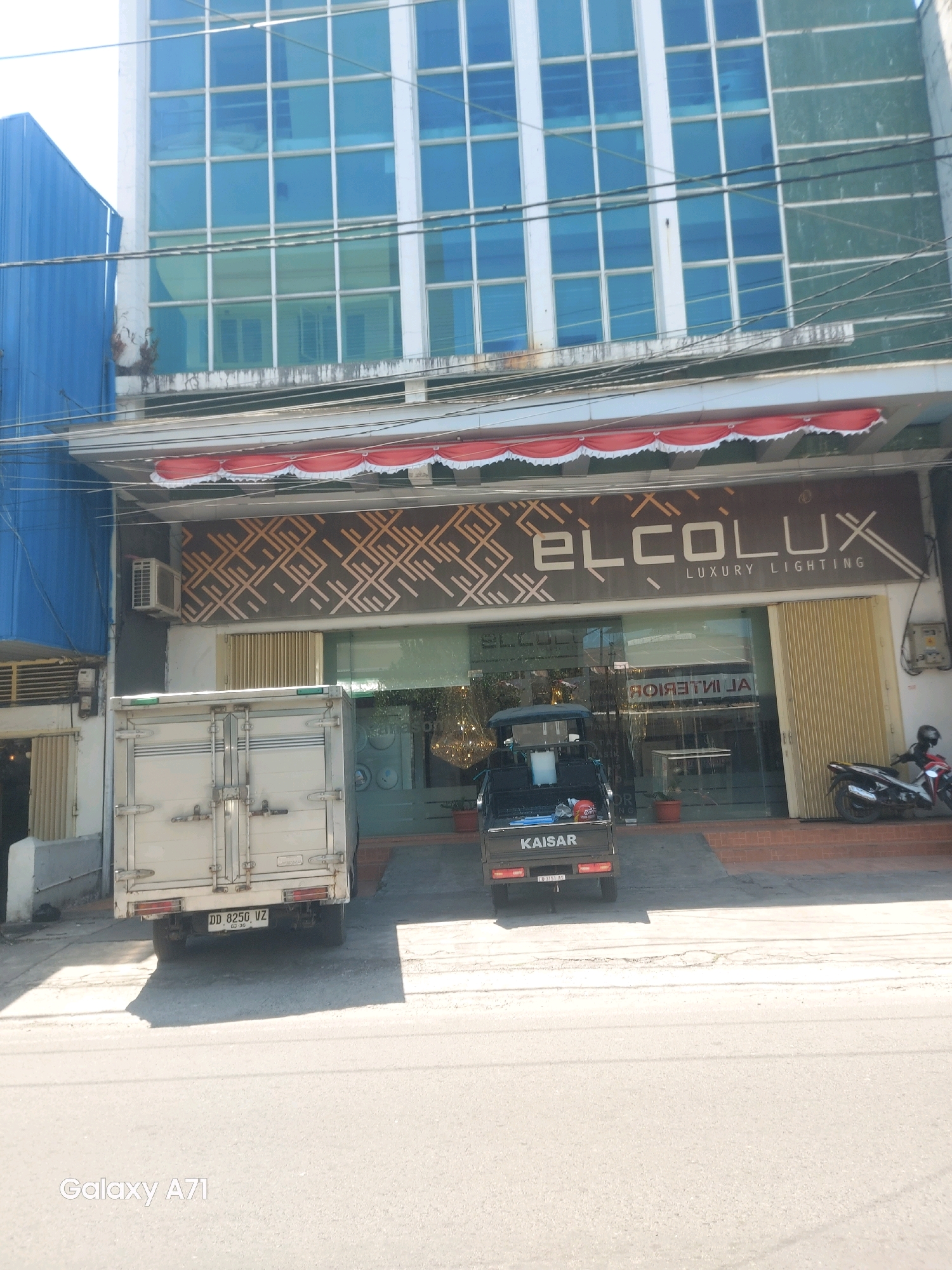 The image shows the exterior of a building in Makassar, Indonesia. The building features a modern facade with large blue tinted windows. A sign above the entrance reads "elCOLUX LUXURY LIGHTING" in a stylized font. Below the sign, there is a glass storefront with vertical blinds on the right side. In front of the building, a white box truck is parked, with a license plate that reads "DD 8250 VZ". To the right of the truck, a small motorized tricycle is parked. A motorcycle is parked near the entrance to the building. The street in front of the building is paved and appears to be a commercial area. The image also includes the "Galaxy A71" watermark, indicating it was taken with a Samsung Galaxy A71 phone.