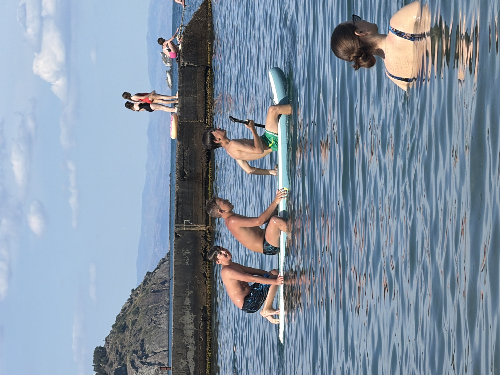 The image depicts a group of people at the sea in Partenit, Ukraine, on a sunny day. In the foreground, a woman with dark hair in a bun, wearing sunglasses and a patterned swimsuit, is partially submerged in the water, facing away from the camera. In the middle ground, three young boys are on a paddleboard. The boy on the left is kneeling and looking back at the camera, while the boy in the middle is also kneeling. The boy on the right is sitting and holding a paddle, wearing green shorts. In the background, a concrete pier extends into the water. On the pier, two young women in swimsuits are standing, and another person is crouched down. Further in the distance, a rocky, tree-covered hill rises from the coast, with mountains visible beyond. The sea is calm, with gentle ripples reflecting the blue sky and scattered clouds.