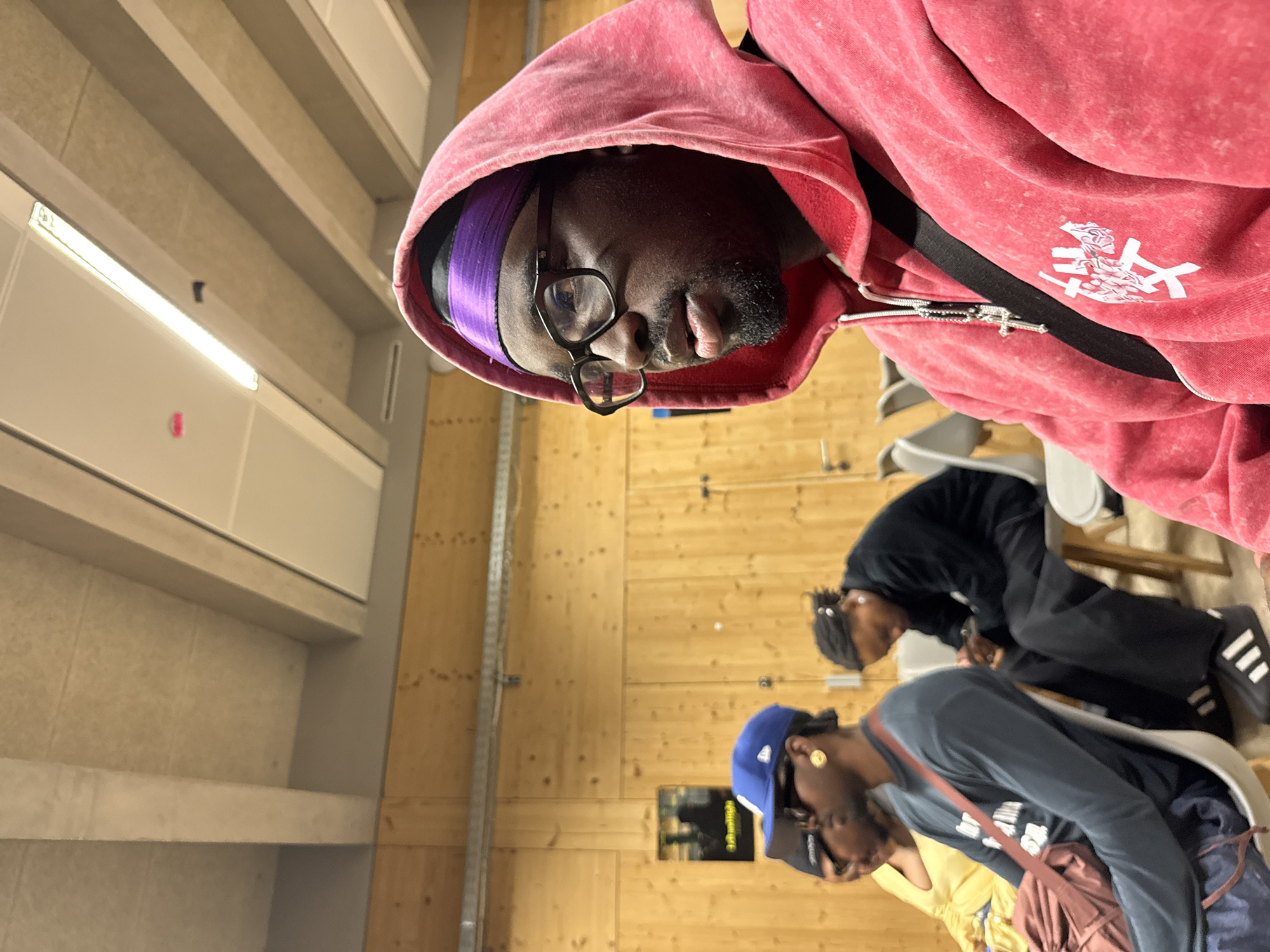 A medium shot depicts a Black male wearing a pink hoodie, glasses, and a purple head covering. He is positioned in the foreground of the image. Behind him, in the middle ground, are two other individuals seated on chairs. One is wearing a blue baseball cap and a grey long-sleeve shirt, while the other is wearing all black. The background reveals a room with light-colored walls and ceiling fixtures. Based on additional information, the location of the scene is within the city of Saint-Ouen-sur-Seine, France. The individuals are interacting in an indoor setting, possibly a classroom or studio.