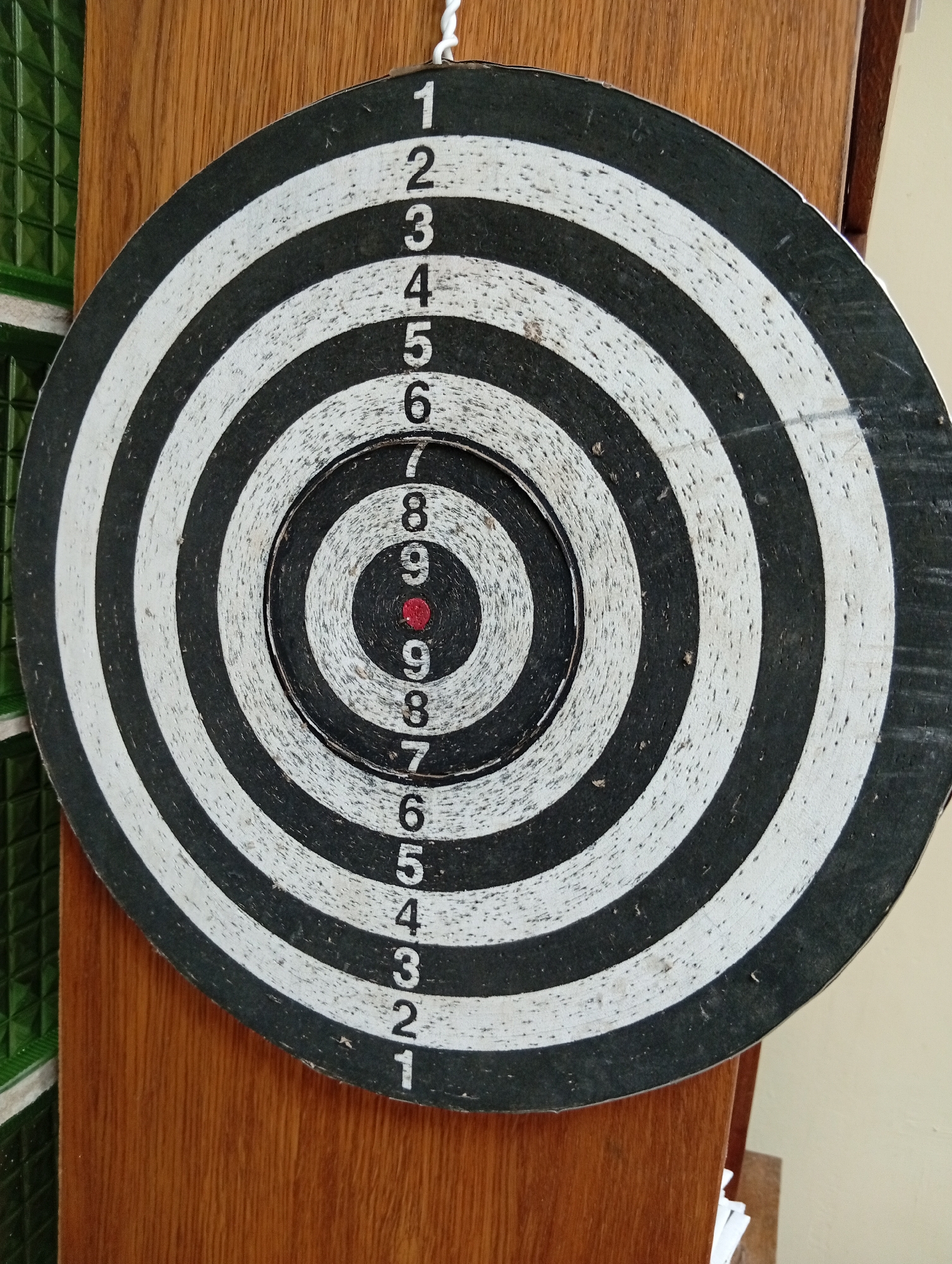 A dartboard is hanging on a wooden surface, possibly a door or wall, with a green tiled surface visible to the left. The dartboard is round and displays concentric rings of black and white. Numbers are printed vertically along the right side of the dartboard, ranging from 1 to 9, with the numbers 9 appearing twice in the center. The bullseye is a small red circle. The dartboard appears worn and slightly damaged.