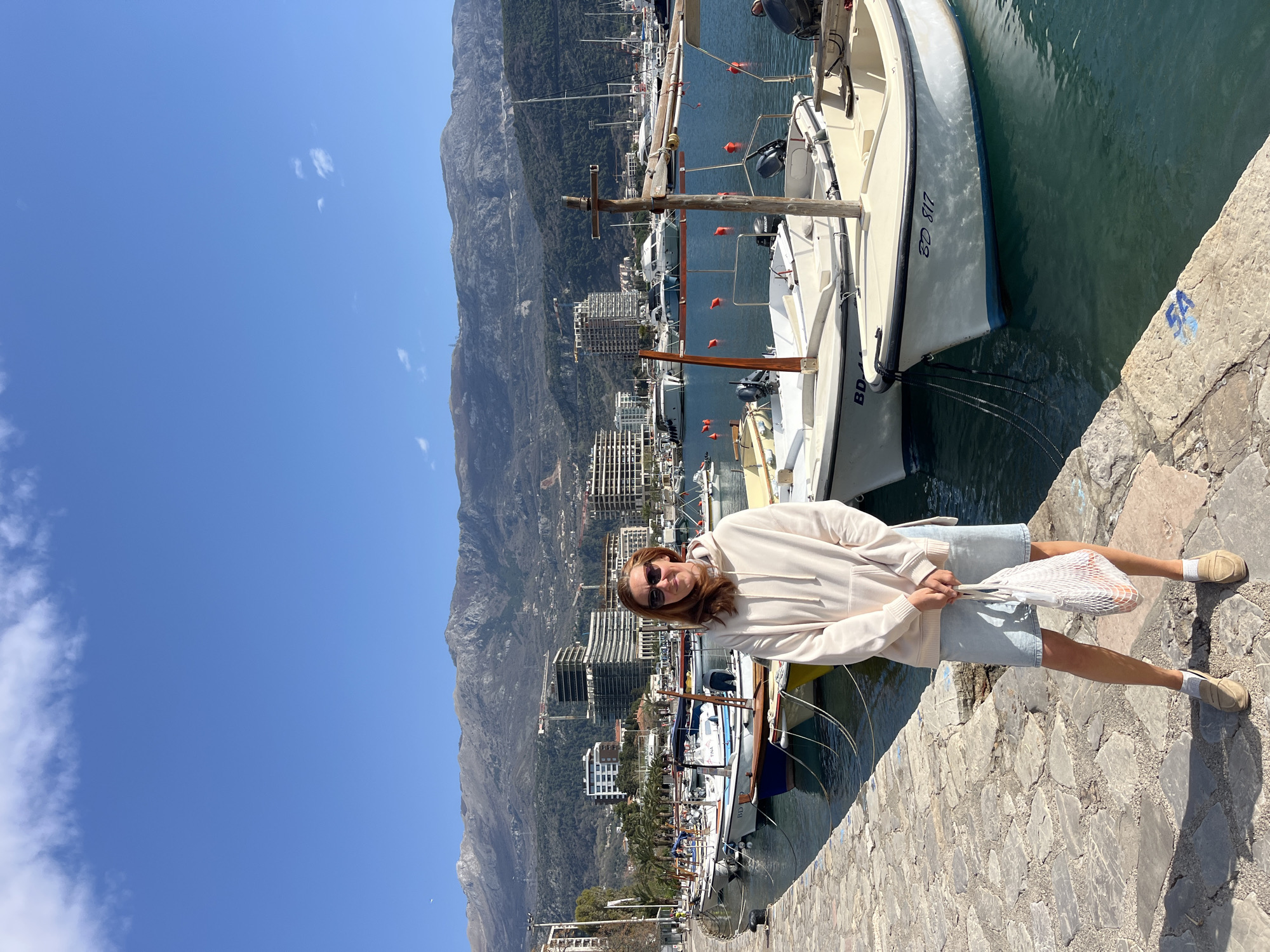 The image shows a person standing on a stone pier in Budva, Montenegro. Mountains are visible in the background, as well as a harbor with several boats. The person is wearing a light-colored hoodie and shorts and is holding a white string bag. The sky is blue with a few clouds.