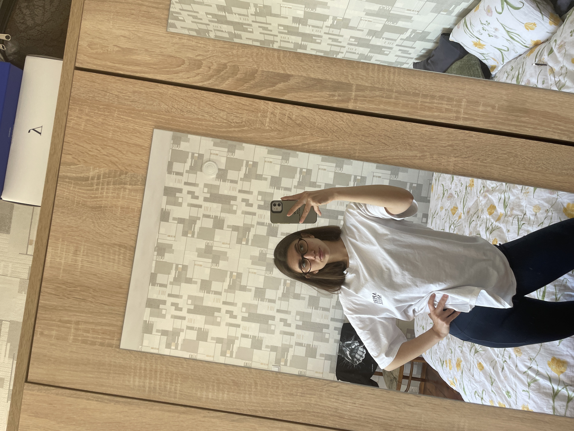 A young woman is taking a mirror selfie in what appears to be a bedroom. The reflection shows a room with a bed covered in floral patterned bedding. The woman has dark hair and is wearing glasses, a white t-shirt, and dark leggings. She is holding a smartphone up to take the picture. The wall behind her is covered in a patterned wallpaper. To the left of the mirror is a wooden wardrobe. Based on the interior decor and the type of buildings likely reflected in a window, it is not possible to specifically determine whether the location is in Sofia, Bulgaria.
