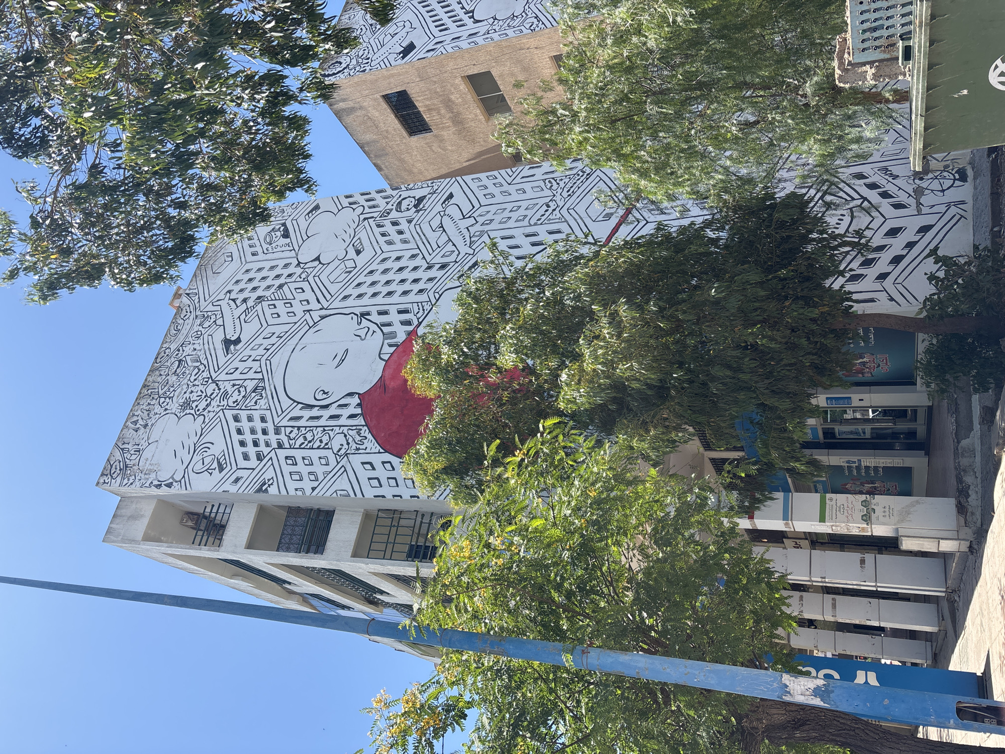 The image shows a mural on the side of a building in a city. The mural is a black and white drawing of a stylized city scene with a large red shape, possibly a cape, draped over a central figure's head. Trees are in the foreground, obscuring parts of the building and the mural. A blue lamppost is on the left side of the image. The building appears to be made of concrete and has multiple stories with windows and balconies. The sky is clear and blue. In the foreground, there are also some shops with signs in Arabic script. A green dumpster is visible on the right. The scene depicts a bright, sunny day in an urban environment.
