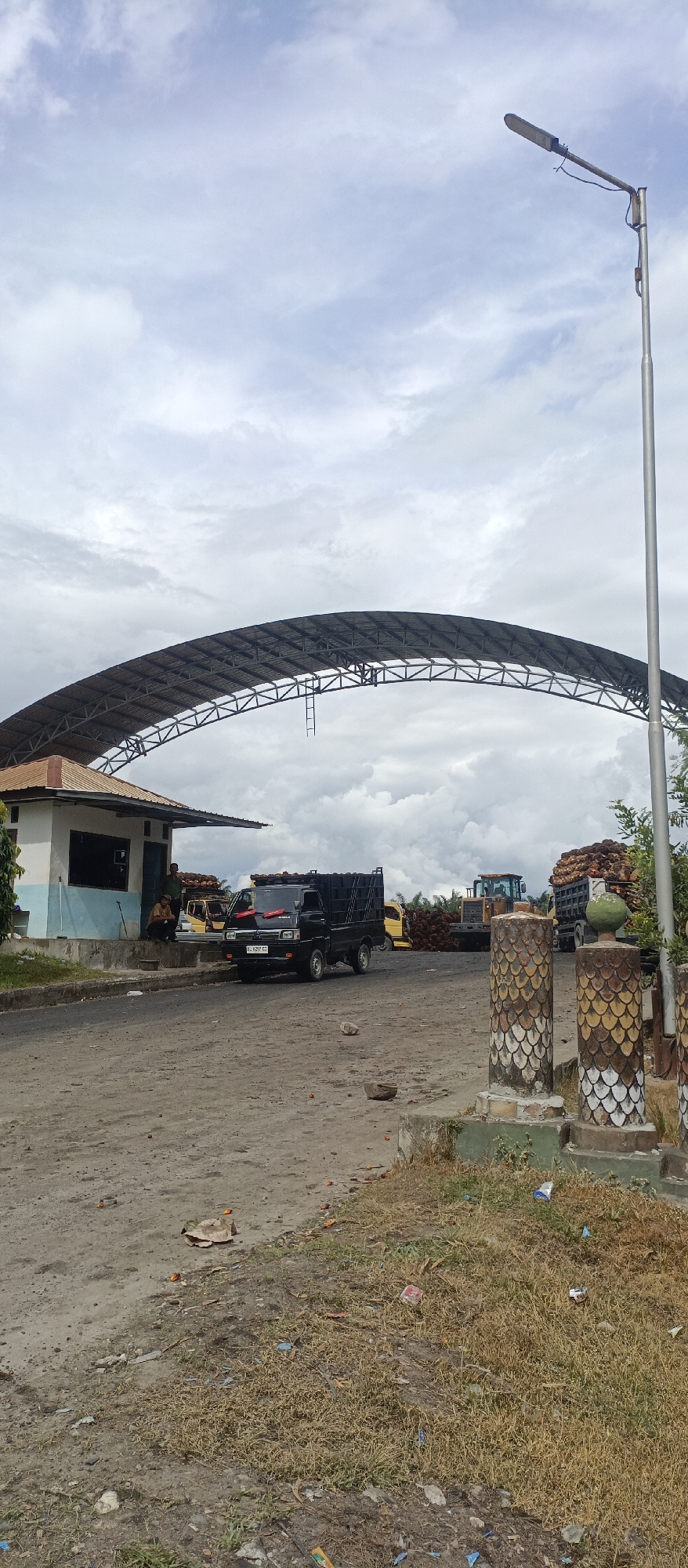 The image depicts a scene at an outdoor facility, possibly a palm oil plantation processing area, in Iemirah, Indonesia. A large, arched metal canopy structure dominates the upper central portion of the image. Underneath the canopy, a small, light blue and white building with a red-tiled roof is visible on the left. Two men are seated near this building. In front of the building, a black pickup truck is parked on a dirt road. Further in the background, a yellow bulldozer and a truck loaded with palm oil bunches are present. The ground is unpaved and dusty, with some scattered debris. The sky is overcast with scattered clouds. Two decorative pillars, featuring a patterned design of scales in yellow, brown, and white, stand prominently on the right side of the foreground. A tall street lamp is positioned on the far right. The overall impression is of an operational area within an industrial or agricultural setting.