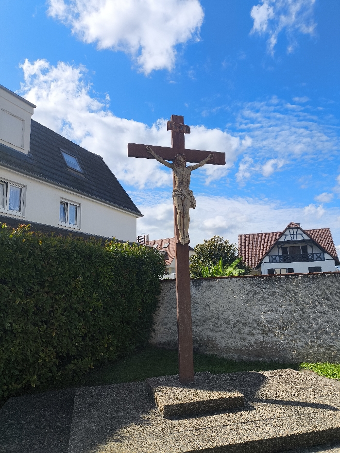 This image depicts a crucifixion monument in Kilstett, France. A weathered, light-colored statue of Jesus Christ is affixed to a prominent, reddish-brown cross, centrally positioned. The cross stands on a multi-tiered, rectangular base constructed from grey, textured aggregate material, with distinct shadows cast upon it by bright sunlight. To the immediate left, a tall, dense green hedge is visible. Behind the hedge and the crucifix, a rough, light-colored stone or concrete wall extends. In the background, two residential buildings are present: a white, multi-story building with a dark tiled roof and multiple rectangular windows to the left, and a traditional Alsatian-style half-timbered house with a red tiled roof and dark timber accents to the right, partially visible above the wall. The sky above is blue with scattered white clouds. No individuals are present.
