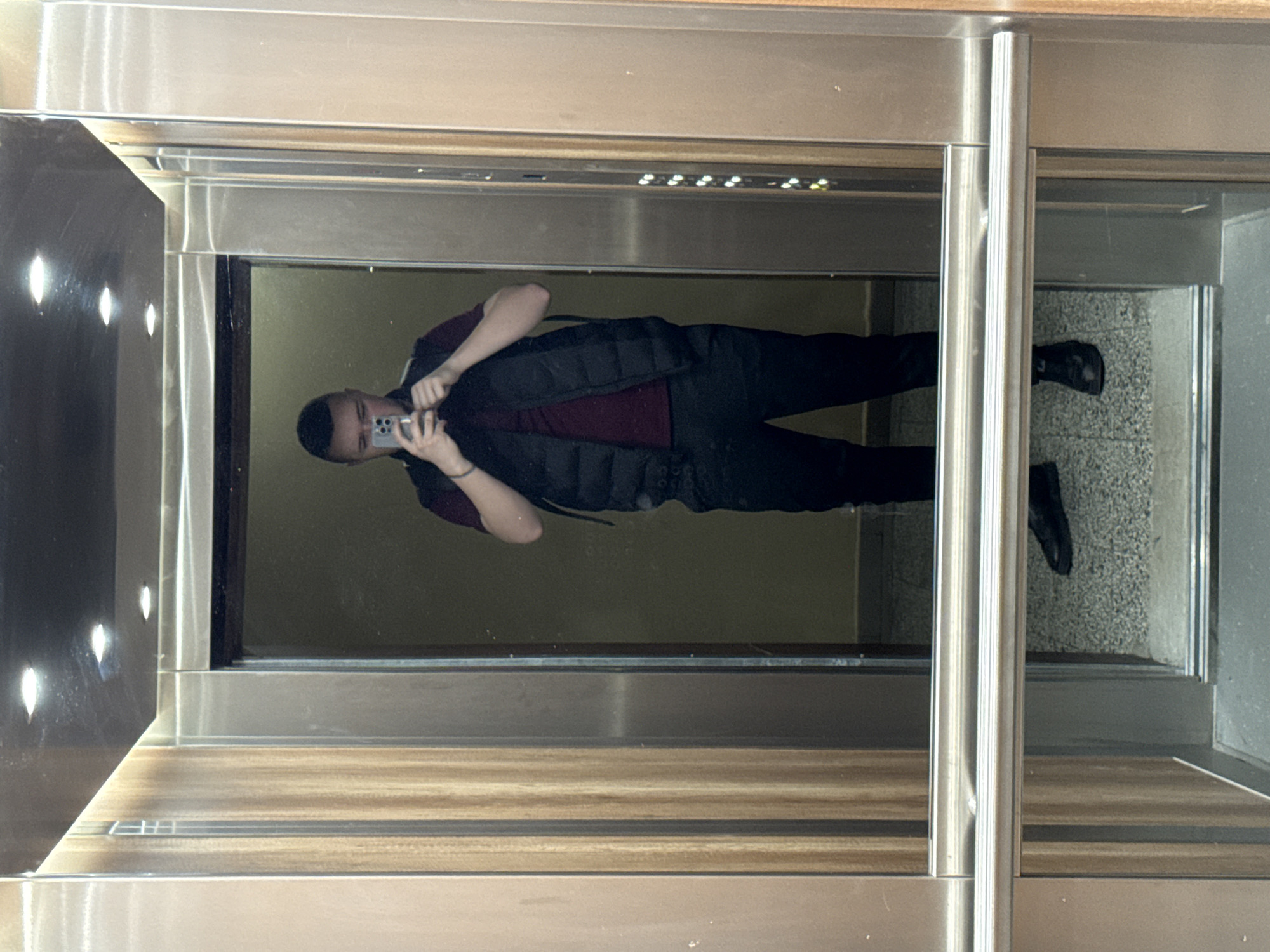 The image is a reflection of a person in an elevator. The person is male, wearing a dark vest over a maroon shirt, and dark pants. He is holding a smartphone, presumably taking a picture of himself in the elevator mirror. The elevator has a modern design with stainless steel and wood-like paneling. The floor of the elevator has a textured grey surface. The location is most likely a building in Sofia, Bulgaria. The elevator doors are partially open, revealing a view of the floor outside.
