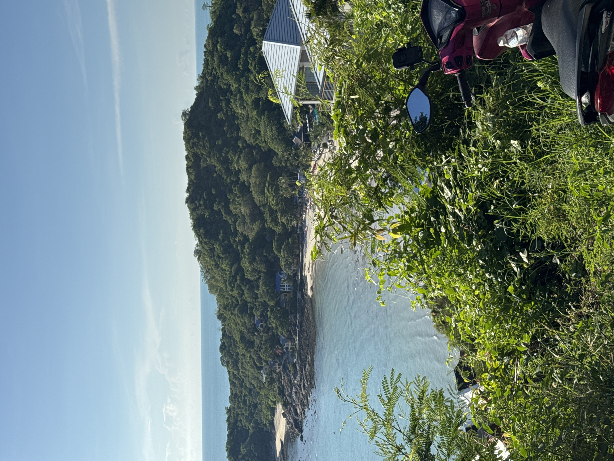 The image displays a vibrant coastal landscape from an elevated viewpoint. In the foreground, lush green foliage and the front portion of a crimson-colored scooter dominate the bottom right corner. The scooter's mirror reflects the clear blue sky. Beyond the vegetation and scooter, a serene bay unfolds, characterized by calm, turquoise waters lapping against a rocky shoreline and a sandy beach. Several small buildings, some with blue roofs, are nestled amongst the trees that blanket the hilly terrain rising from the bay. Further into the distance, the deep blue ocean meets a bright, cloud-dappled sky. The overall scene suggests a tropical or Mediterranean setting, possibly a resort area given the presence of houses and the proximity to the water.